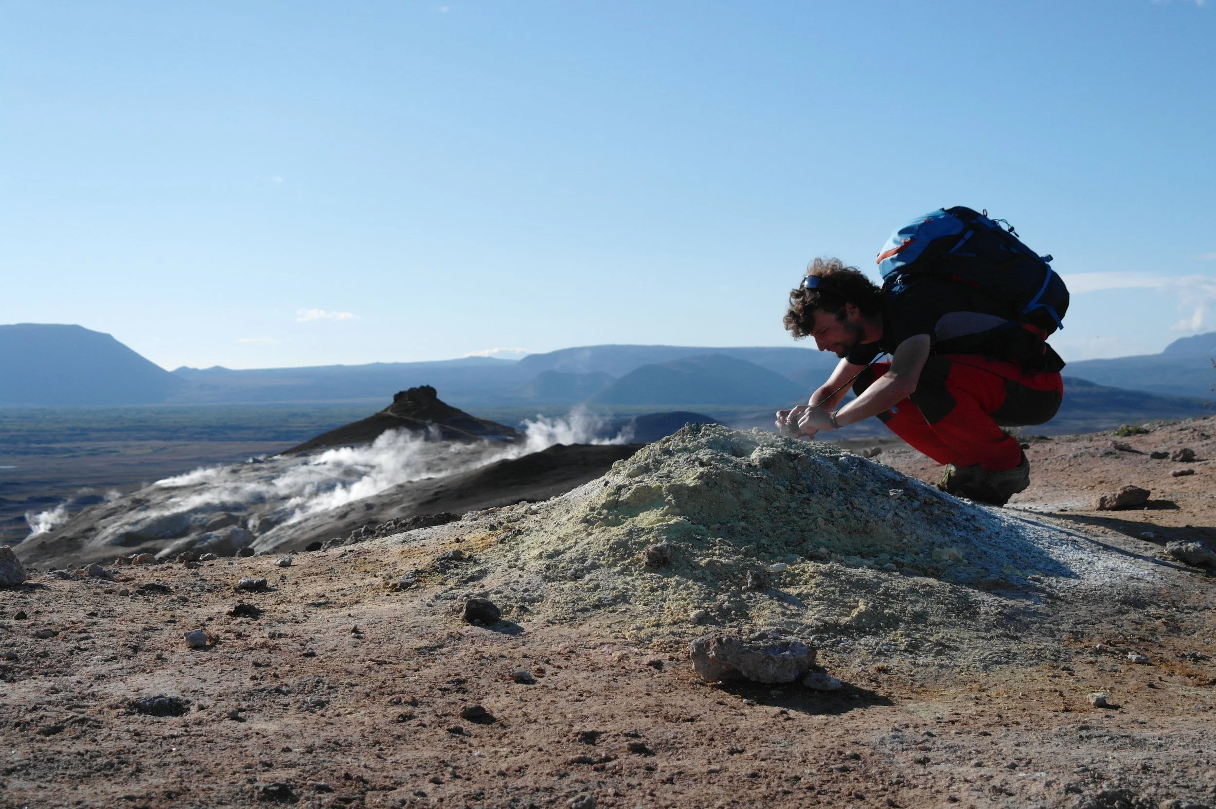 A man crouching in a volcanic landscape with steam vents, wearing a backpack and outdoor gear, surrounded by distant mountains and a blue sky.