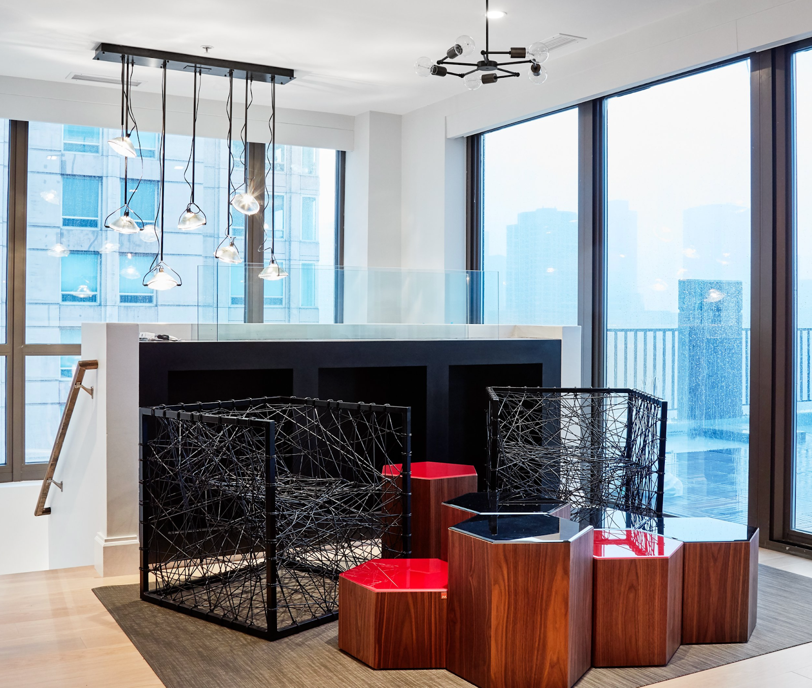 Contemporary lounge featuring black wire cube armchairs, tiered red and wood hexagonal nesting tables, and industrial multi-pendant lighting against floor-to-ceiling city view windows