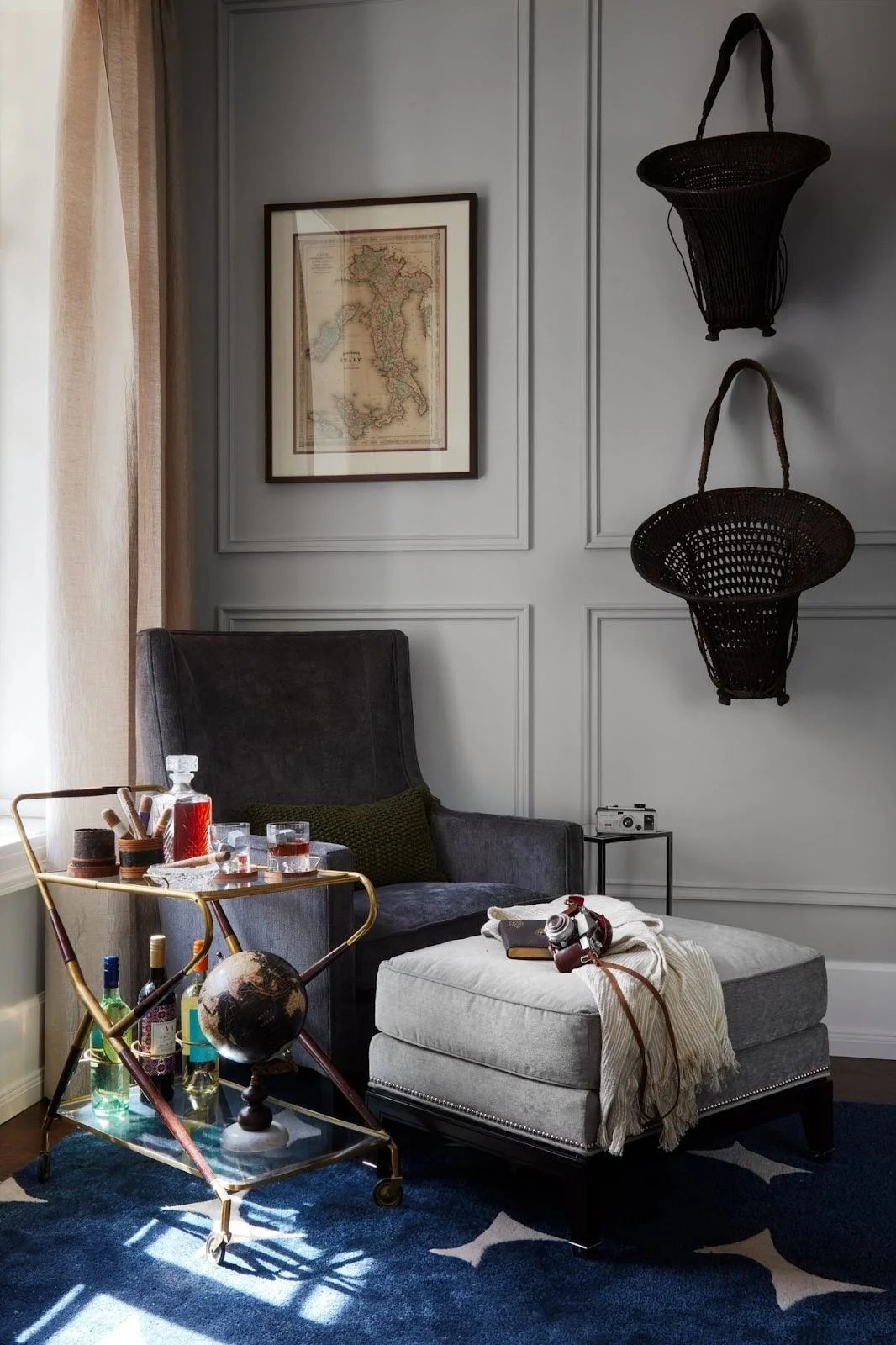 A masculine reading nook featuring a dark grey velvet wingback chair, a grey ottoman, and a gold bar cart against light grey wall molding.