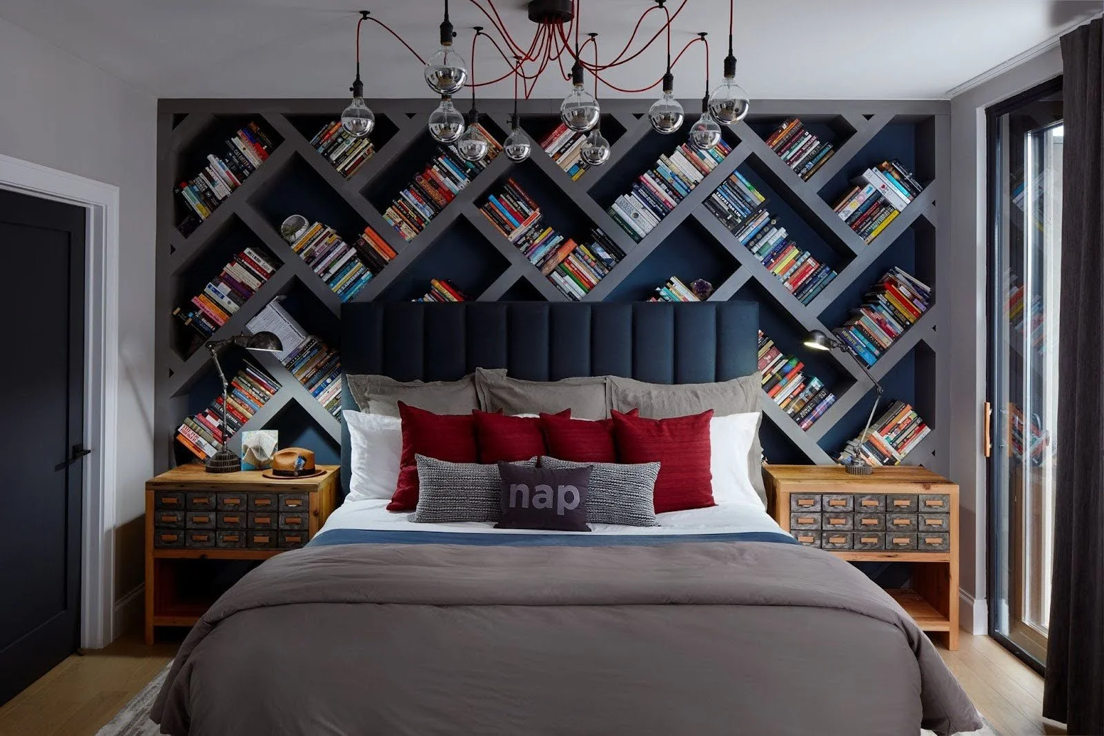 Custom grey diamond-grid bookshelf wall behind a dark upholstered bed with red accent pillows and vintage-style industrial lighting.