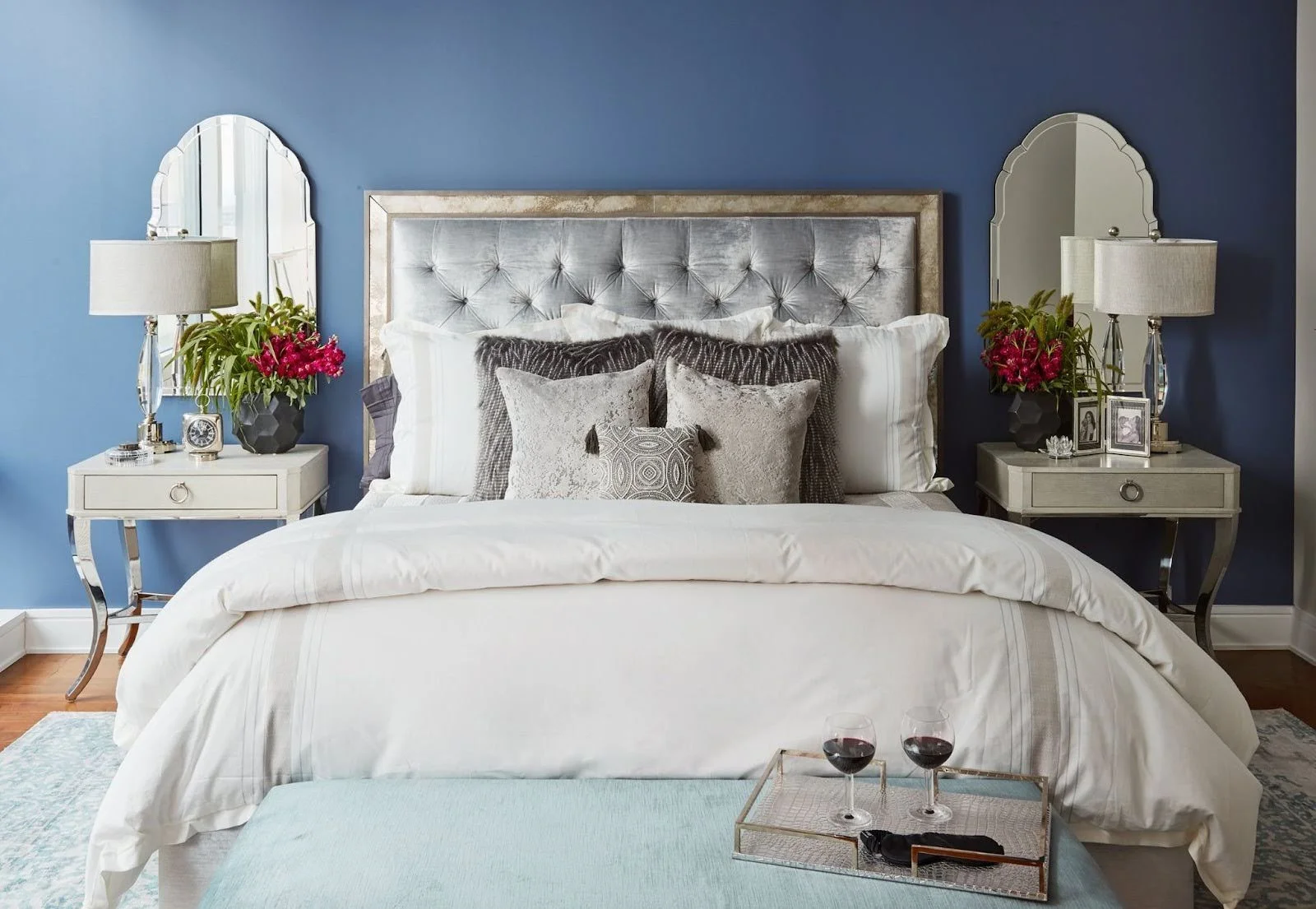 Luxury bedroom with a navy blue accent wall, a silver tufted velvet headboard, white bedding, and symmetrical white nightstands with scalloped mirrors.;