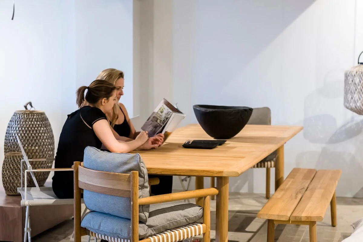 Emily Mackie chicago designer - Two women sit at a light wooden dining table, reading a magazine. The setting feels serene and modern with woven basket decor and a large black bowl centerpiece.