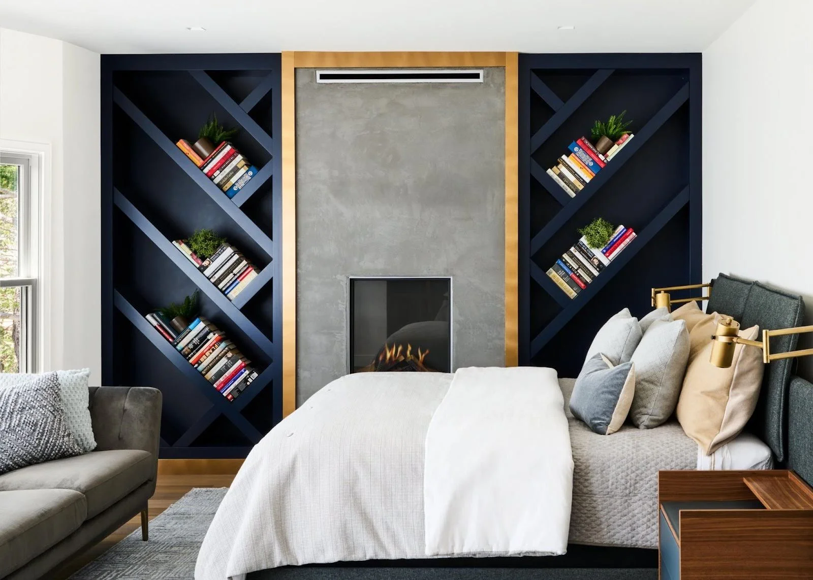 Custom navy blue diagonal bookshelves flanking a modern grey fireplace in a master bedroom with a white bed and gold reading lamps.