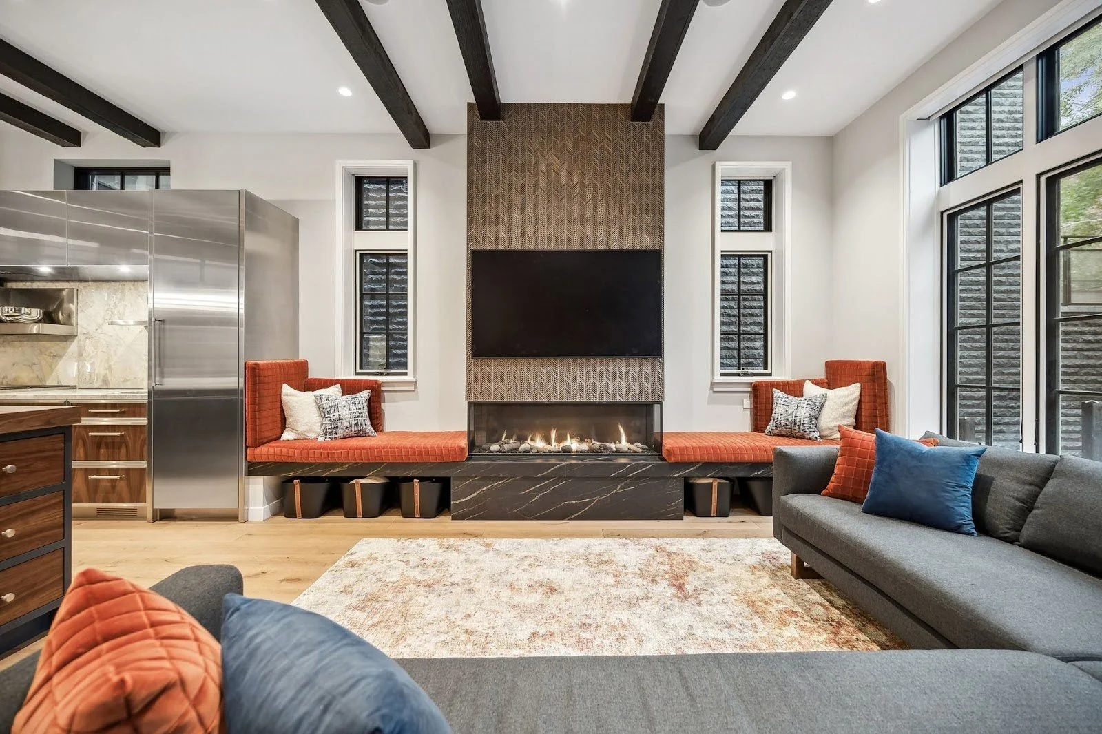 Large open-concept living room in Lincoln Park featuring dark wood ceiling beams, an industrial kitchen with stainless steel appliances, and a central linear fireplace flanked by orange banquette seating.