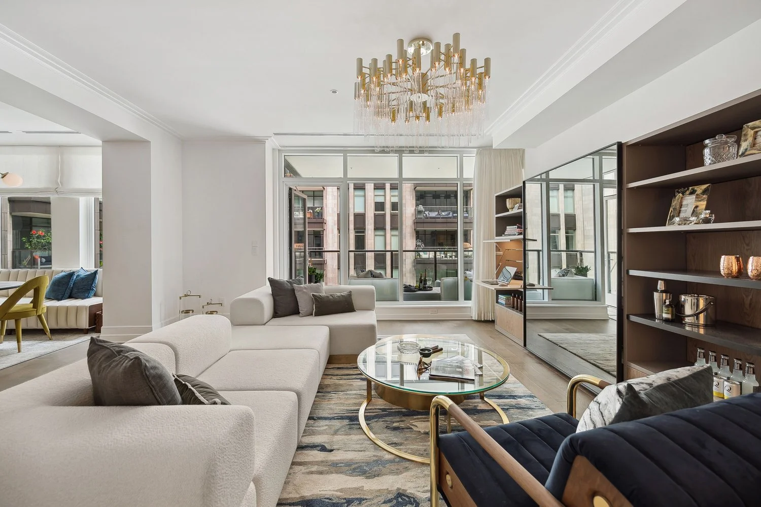 Luxury modern living room featuring a large white sectional, gold statement chandelier, and floor-to-ceiling windows overlooking an urban cityscape
