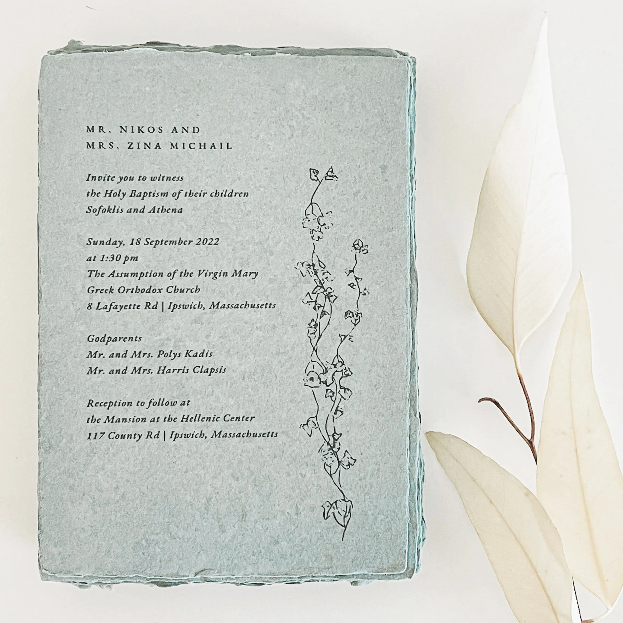 Invitation card for a christening or baptism ceremony with a simple black and white line drawing of a flowering vine on a light blue background, placed on cream-colored paper with dried white leaves beside it.