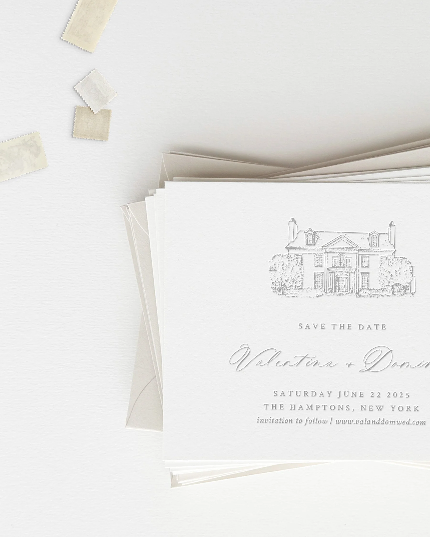 Elegant white wedding save the date card with a house illustration and script text, placed on a white surface with small beige stamps nearby.