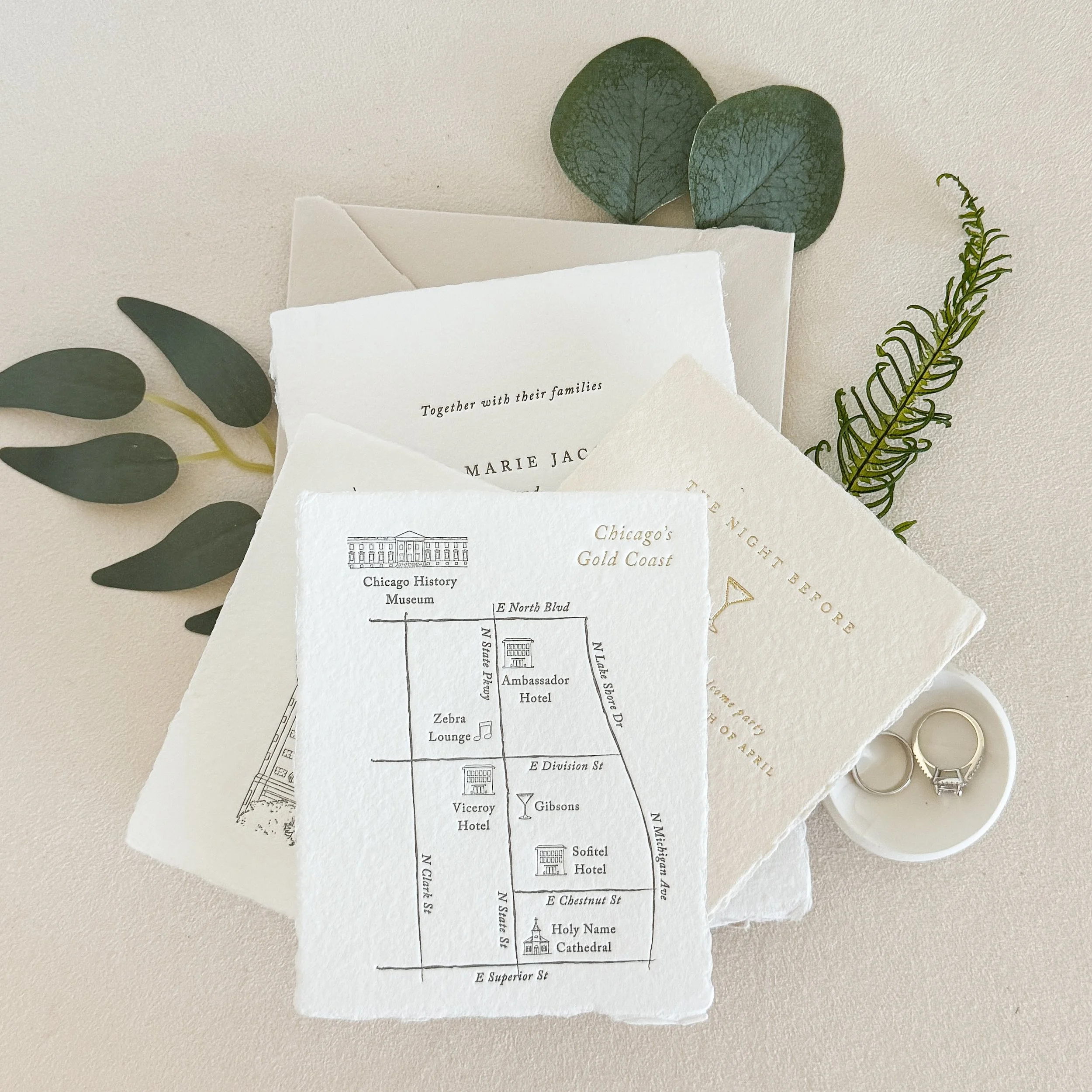 Wedding invitation, greenery, and engagement rings arranged on a light background.