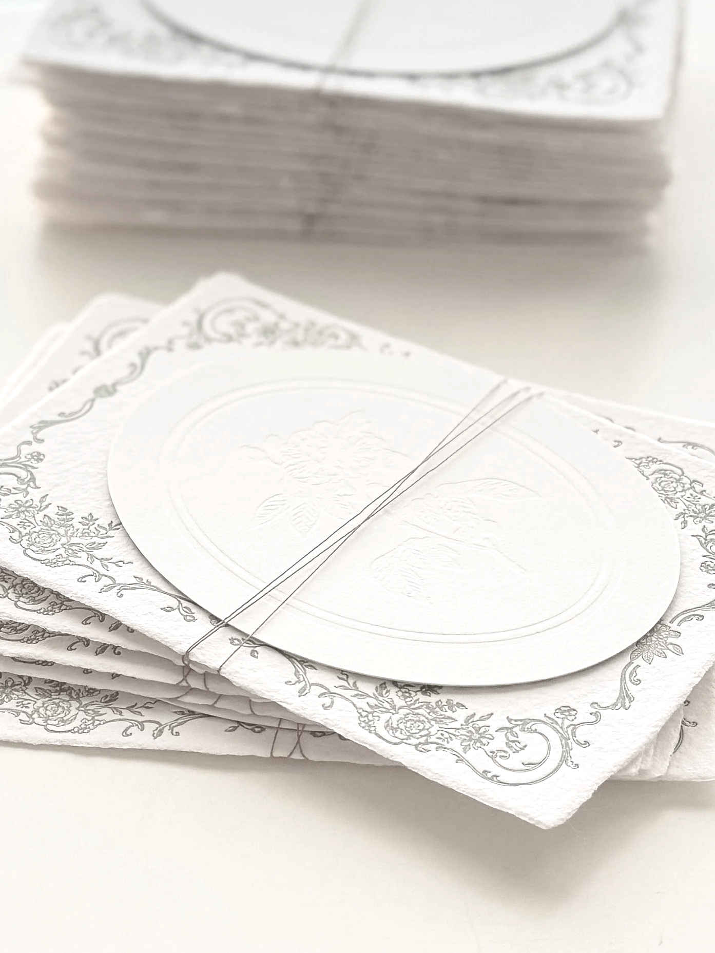 Stack of embossed handmade paper with a decorative border and a blind embossed floral design, with a smaller cut-out oval card on top, placed on a white surface.