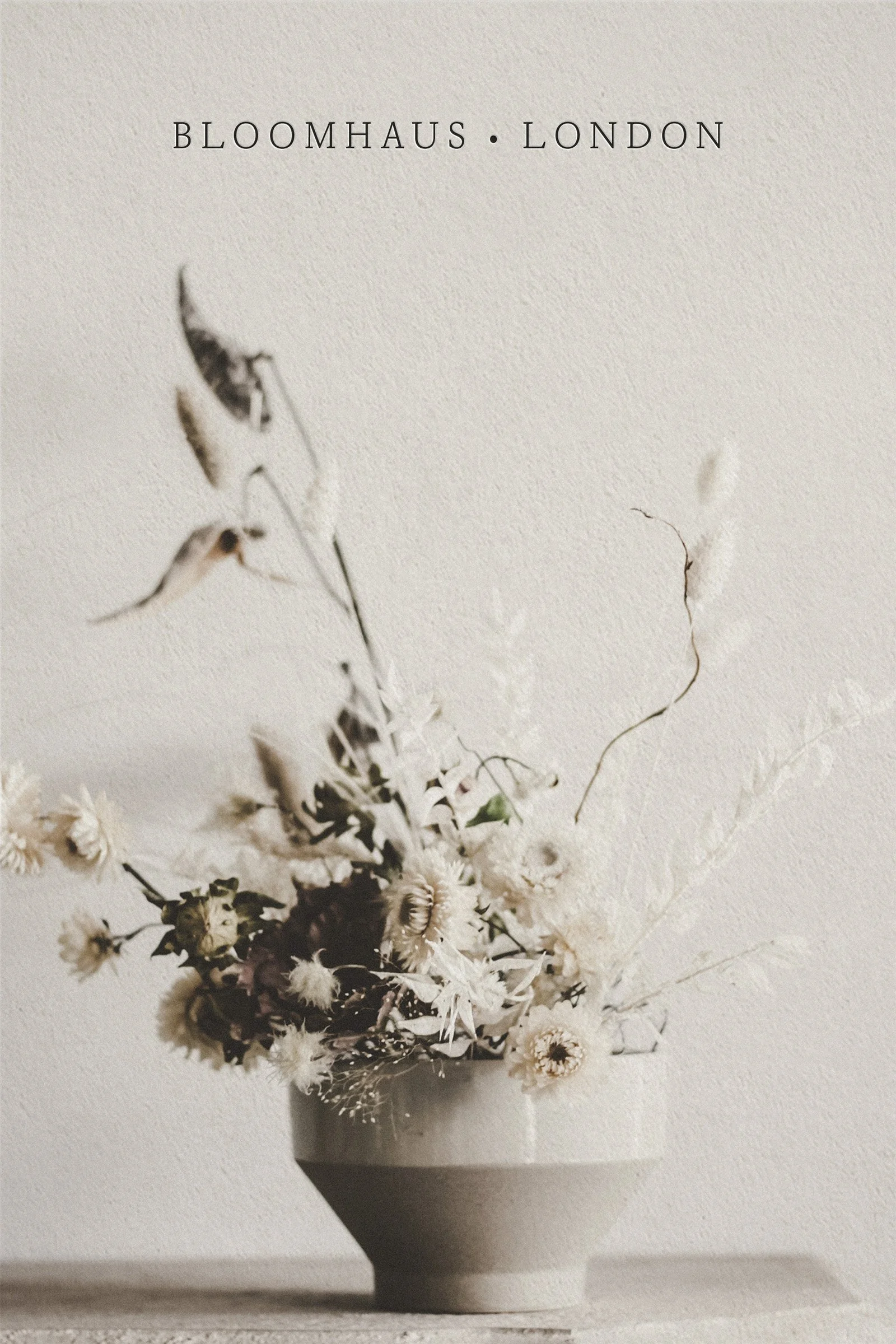 A minimalist image of a beige ceramic vase with dried white flowers on a neutral background. The text 'BLOOMHAUS · LONDON' is printed at the top.