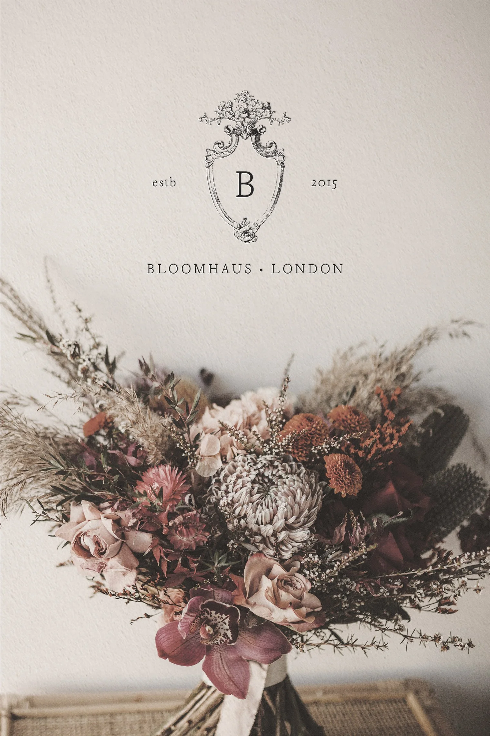 A bouquet of dried and fresh flowers in pink, burgundy, and beige tones, placed in front of a wall art with an ornate crest and text reading 'estb 2015 BLOOMAUS LONDON'.