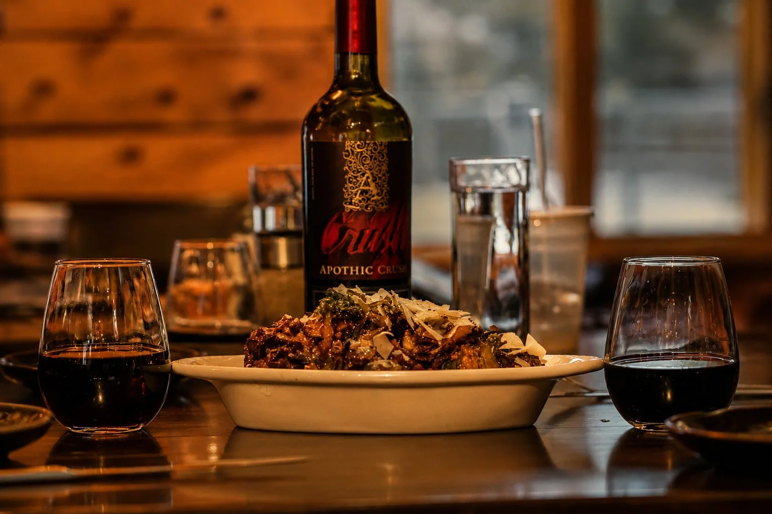 A dinner table with a large white dish of pasta, two glasses of red wine, a bottle of red wine, and several glasses and water bottles in the background, set in a cozy wooden restaurant.