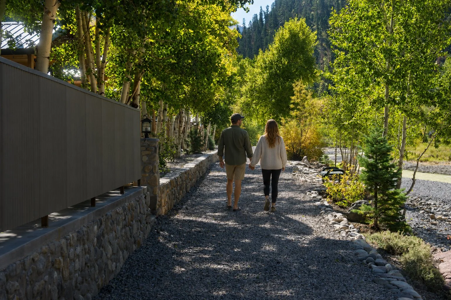 A couple walking hand in hand along a rocky pathway lined with green trees and small plants, with mountains in the background.