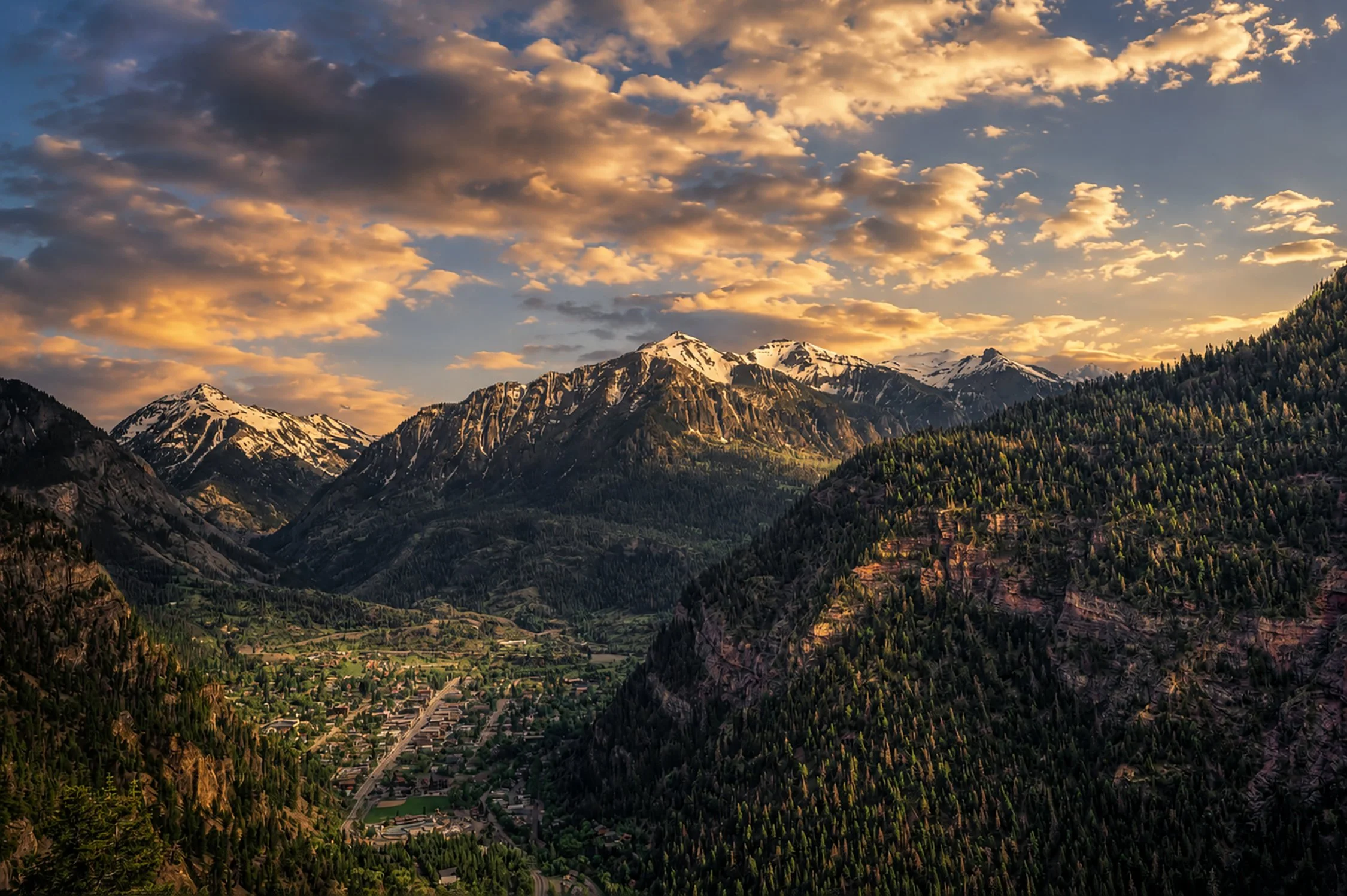 Mountain range with snow-capped peaks under a cloudy sky at sunset, overlooking a valley with a small town surrounded by forests.