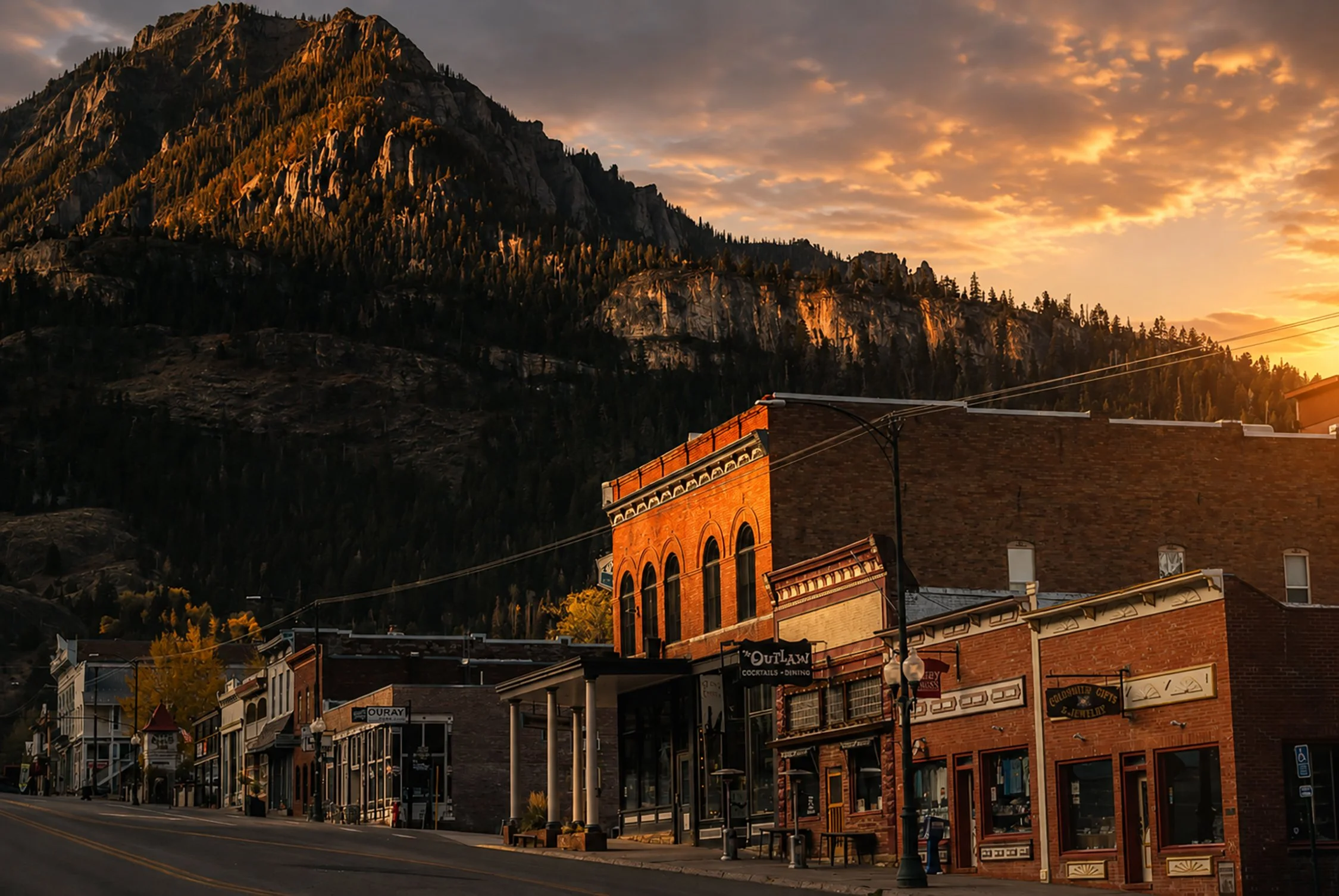 A small town with historic brick buildings along a street, with a mountain covered in trees in the background and a partly cloudy sky during sunset.