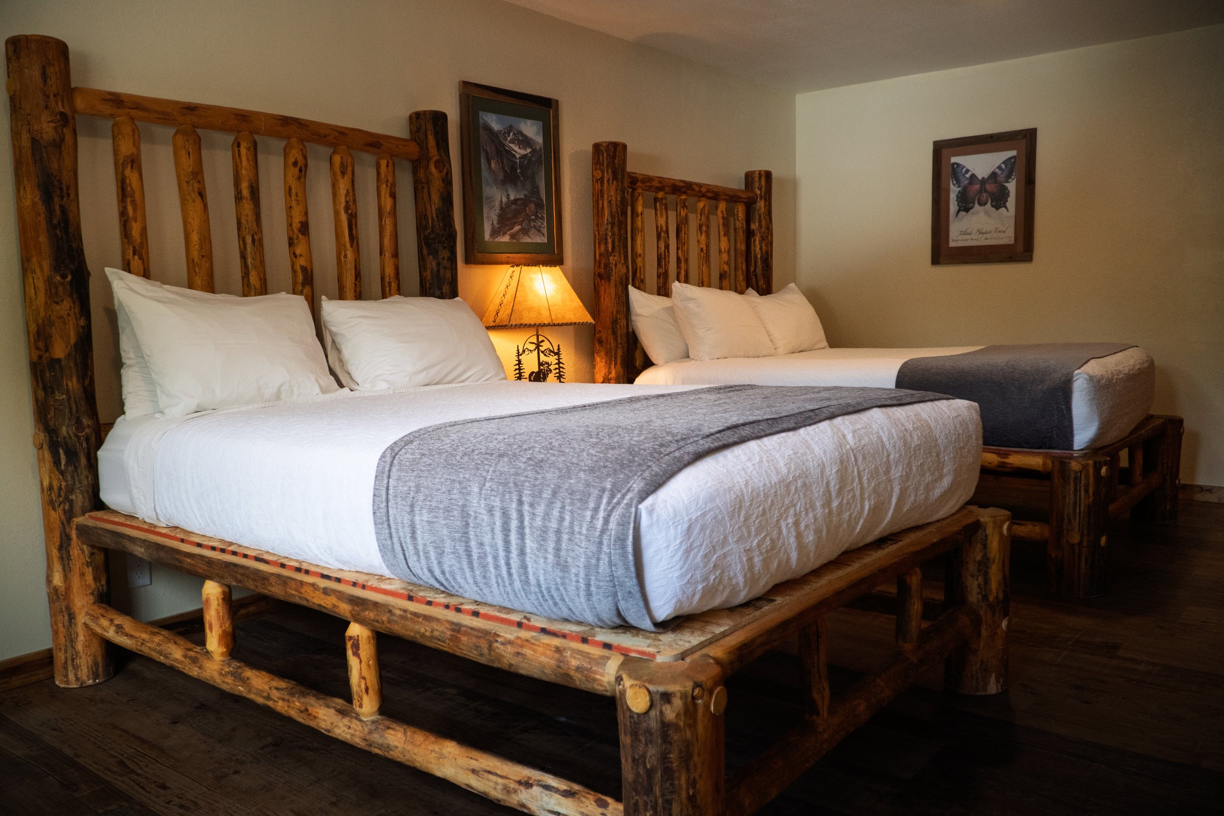 Rustic bedroom with two large log beds, white bedding, and pillows. A nightstand with a lamp featuring a nature design is between the beds. Framed mountain and butterfly pictures hang on the wall.