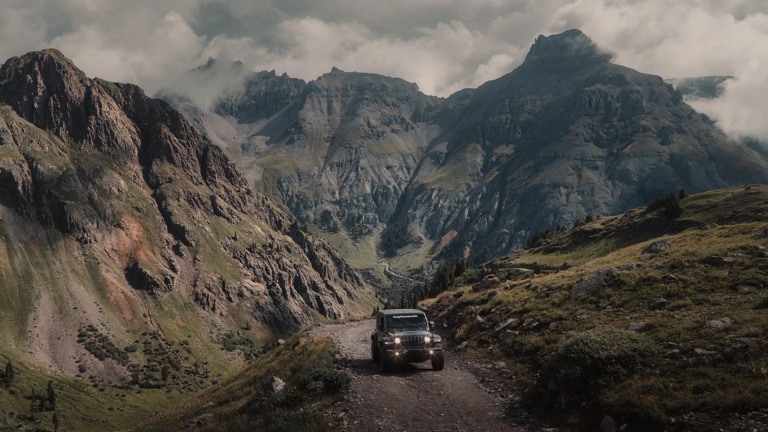 A black Jeep driving on a rugged mountain road surrounded by tall, rocky, green mountains with clouds in the sky.