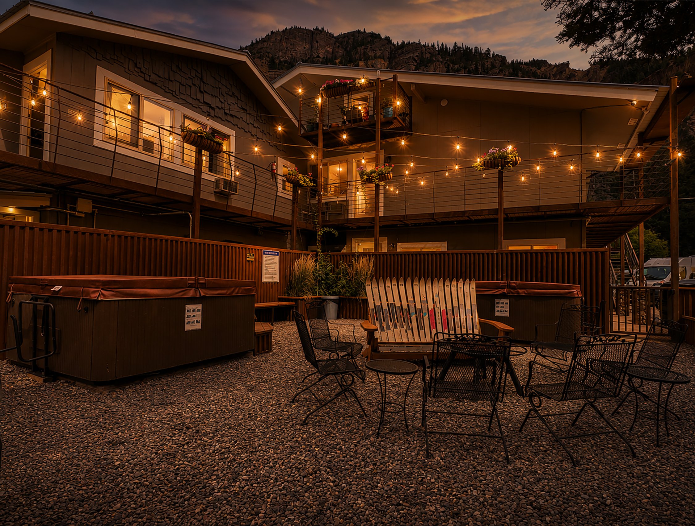 A house with multiple outdoor string lights hanging around a balcony and patio area during dusk. The house has even siding, large windows, and a small balcony with potted plants. There is outdoor furniture and a hot tub on the patio, with a neighboring but darker-skinned mountain or hillside in the background.