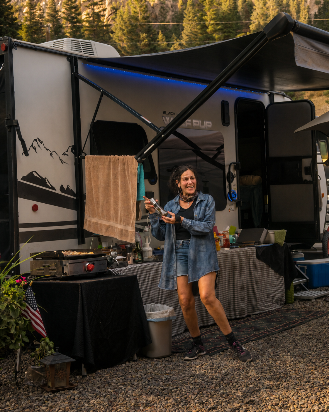 A woman in a denim jacket and shorts is smiling and holding a bottle in front of a camper trailer with outdoor cooking and camping setup amid trees.