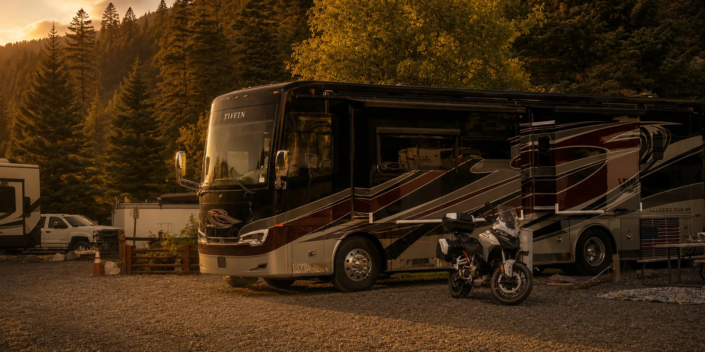 A black and red Tiffin luxury motorhome parked in a campground with tall pine trees and mountains in the background during sunset.
