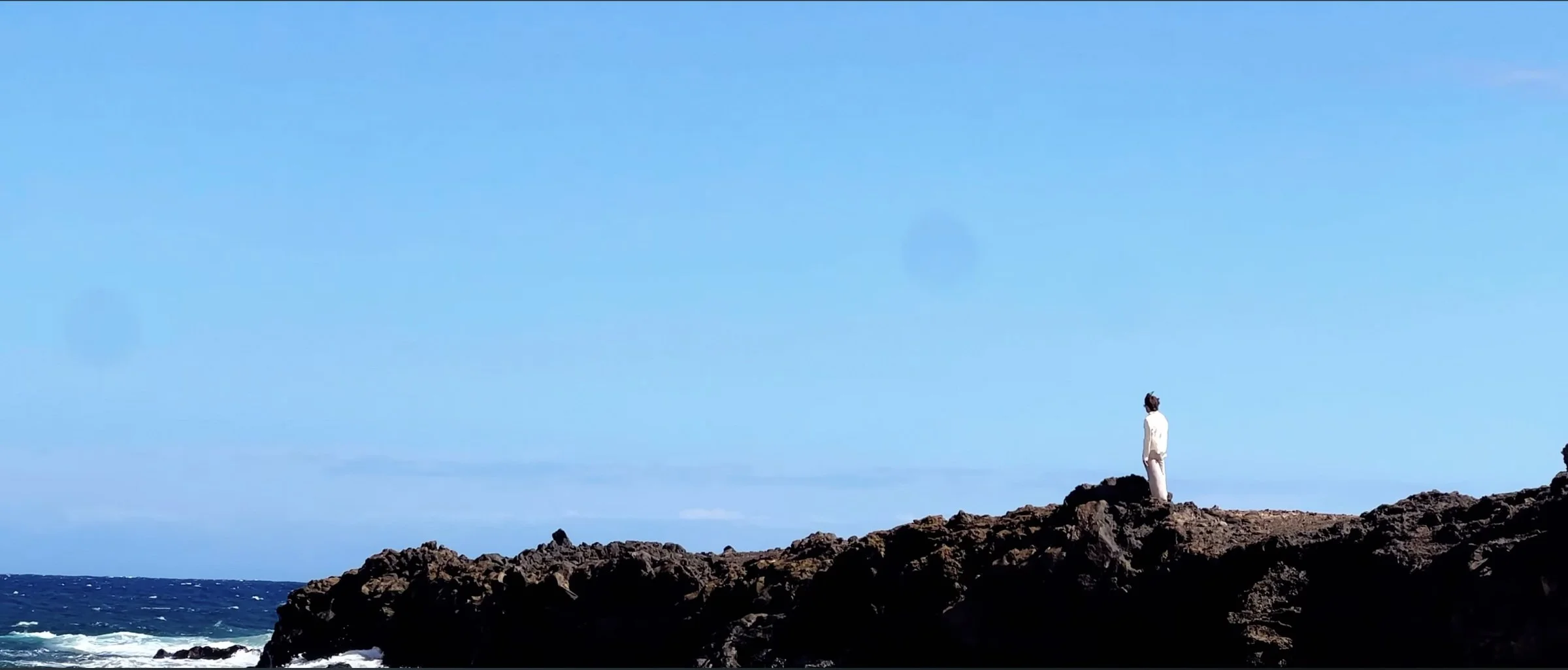 DIUCE vêtu de blanc sur une crête de rochers noirs face à la mer, sous un grand ciel bleu.