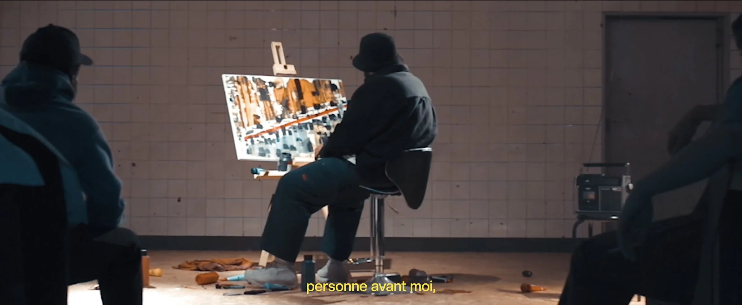 Photogramme de SZERO : travail sur une peinture dans un espace intérieur, entouré de personnes assises.