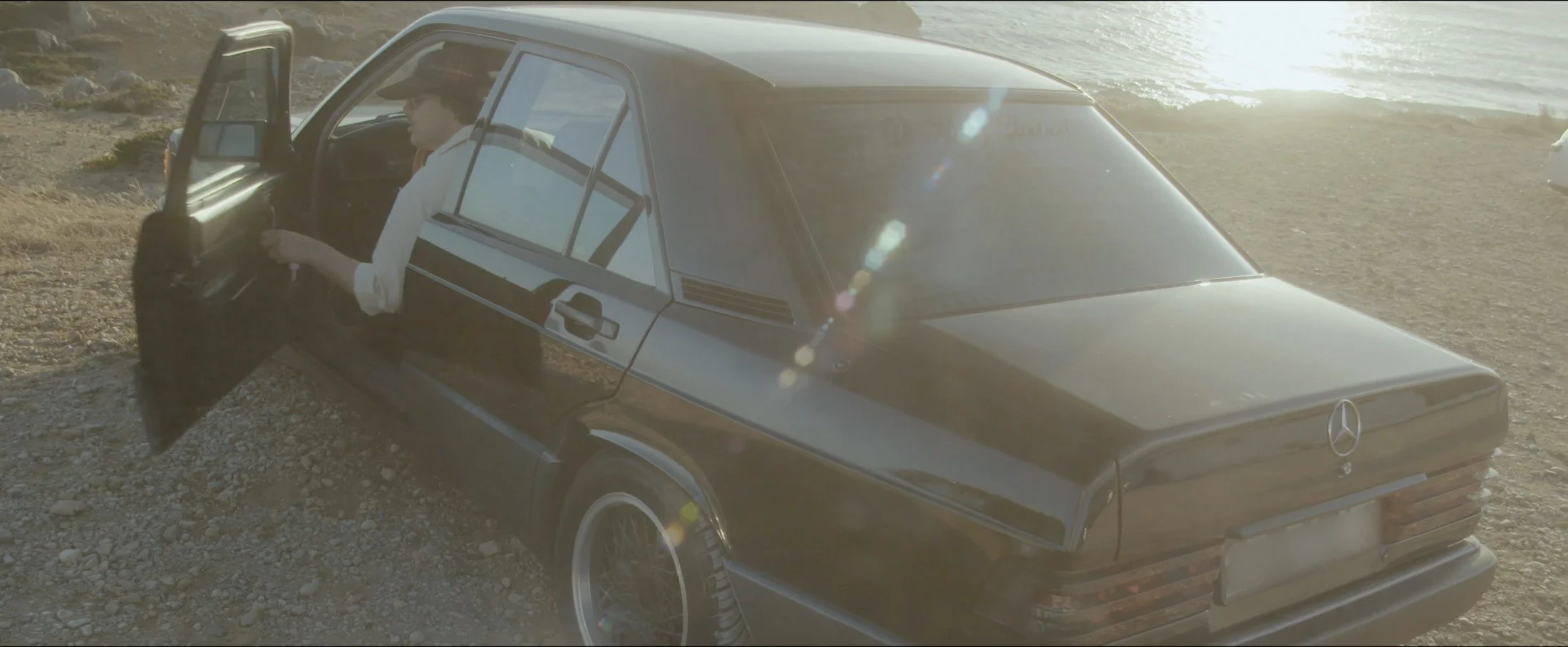 Photogramme de SZERO : une Mercedes noire près de l’eau, éclairée en contre-jour, avec un homme à la portière.