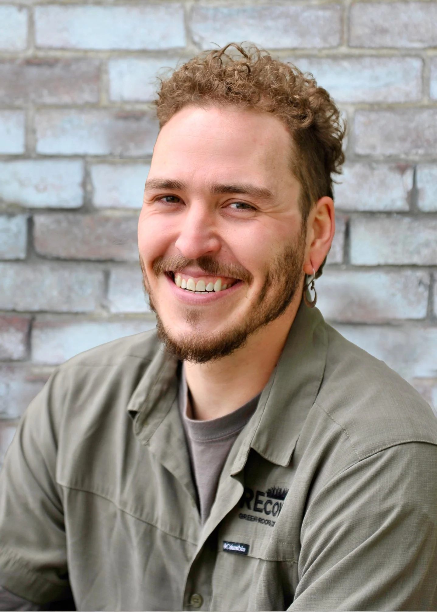 A smiling man with curly hair and a beard, wearing a uniform with a badge, sitting in front of a brick wall.