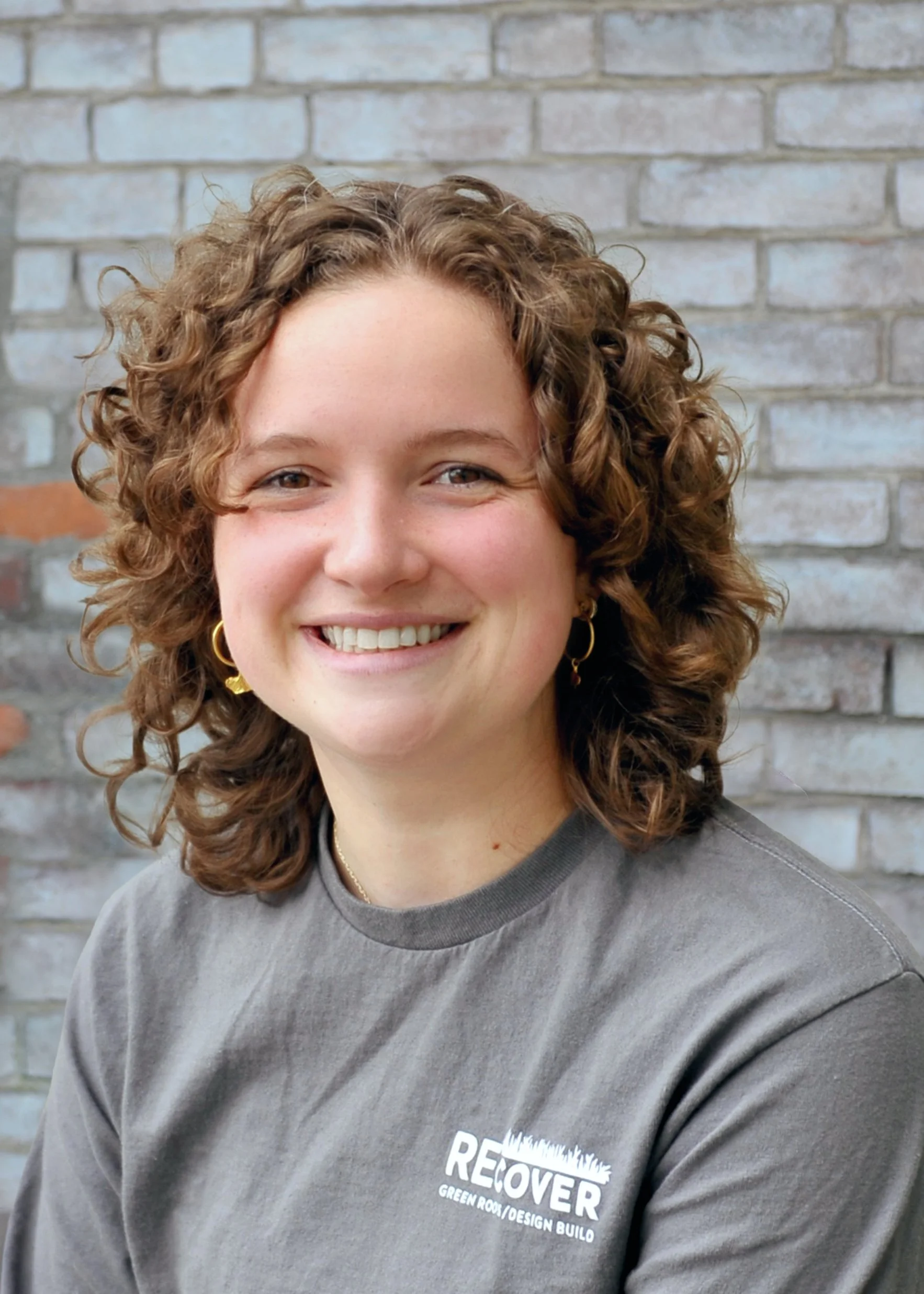 Close-up of a smiling woman with curly hair wearing a gray shirt with a logo that reads 'RE:OVER GREEN ROOF / DESIGN BUILD' against a brick wall background.