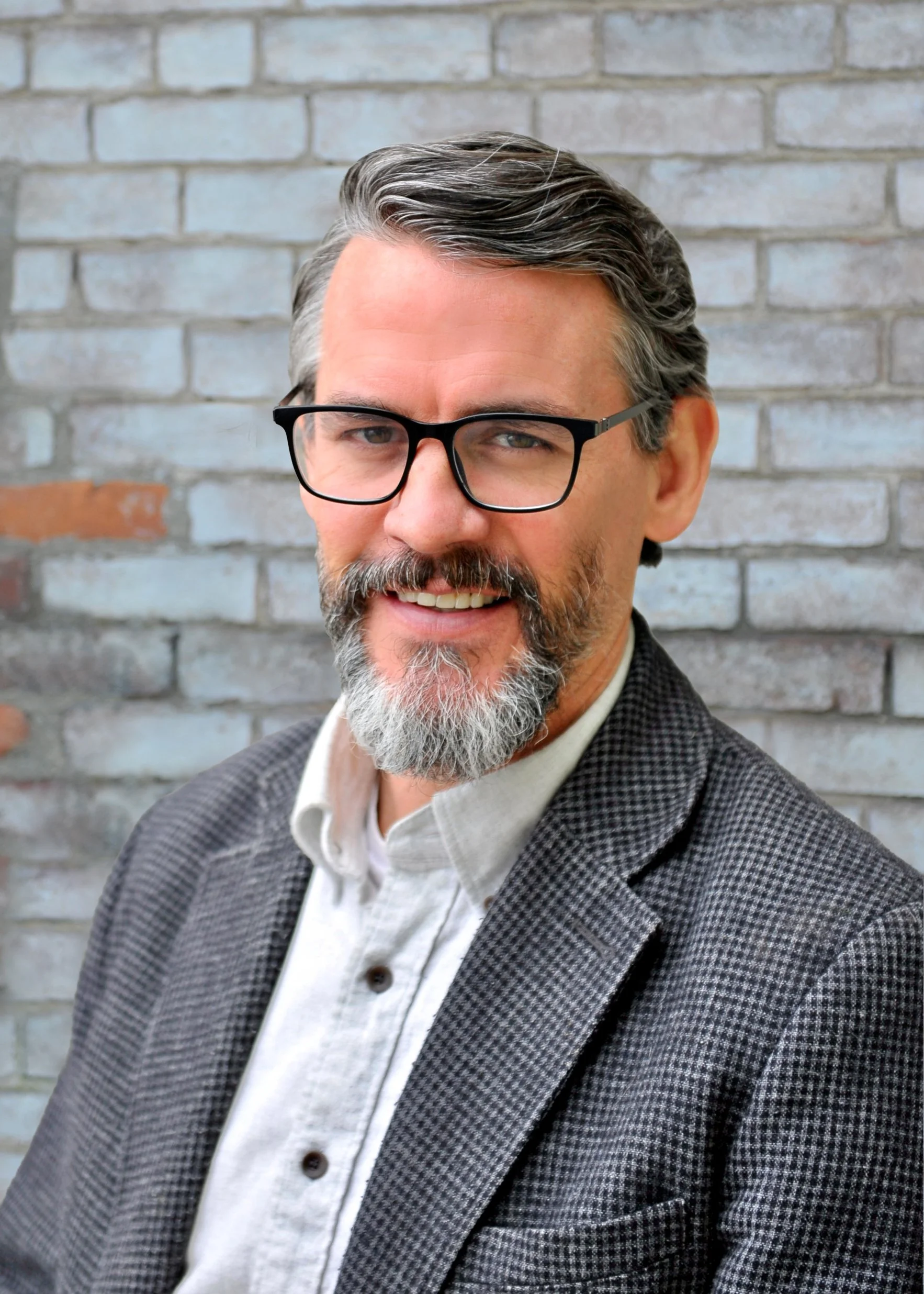Close-up photo of a middle-aged man with gray hair and beard, wearing black-rimmed glasses, a white shirt, and a gray checkered blazer, standing in front of a brick wall.
