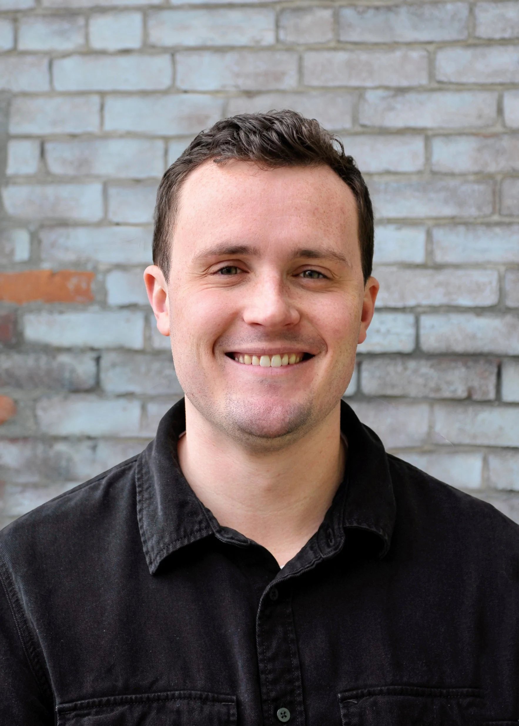 Close-up photo of a smiling young man with short, dark hair, wearing a black jacket, standing in front of a brick wall.