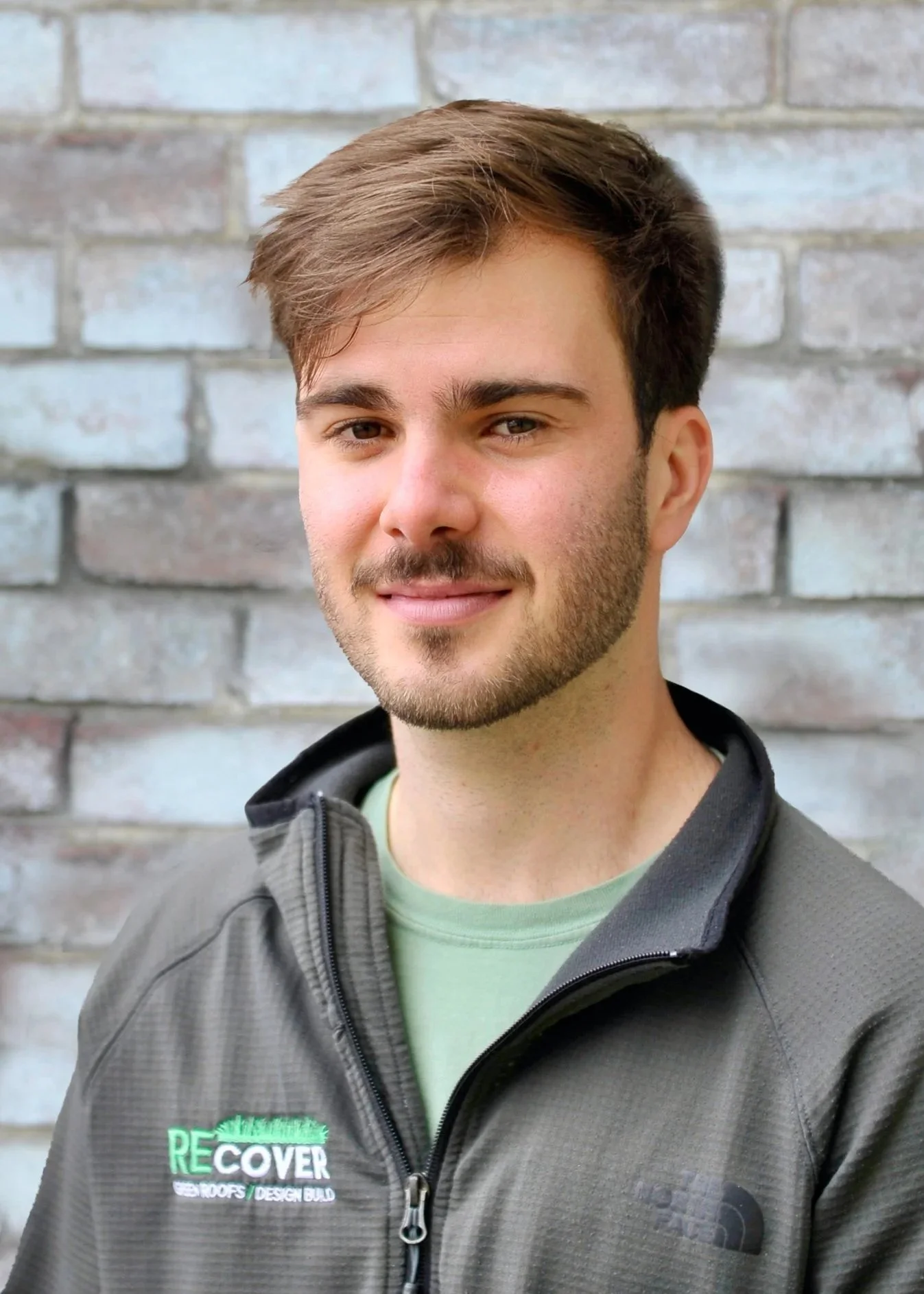 Portrait of a young man with brown hair and a slight beard, wearing a grey jacket with a logo on it, standing in front of a brick wall.