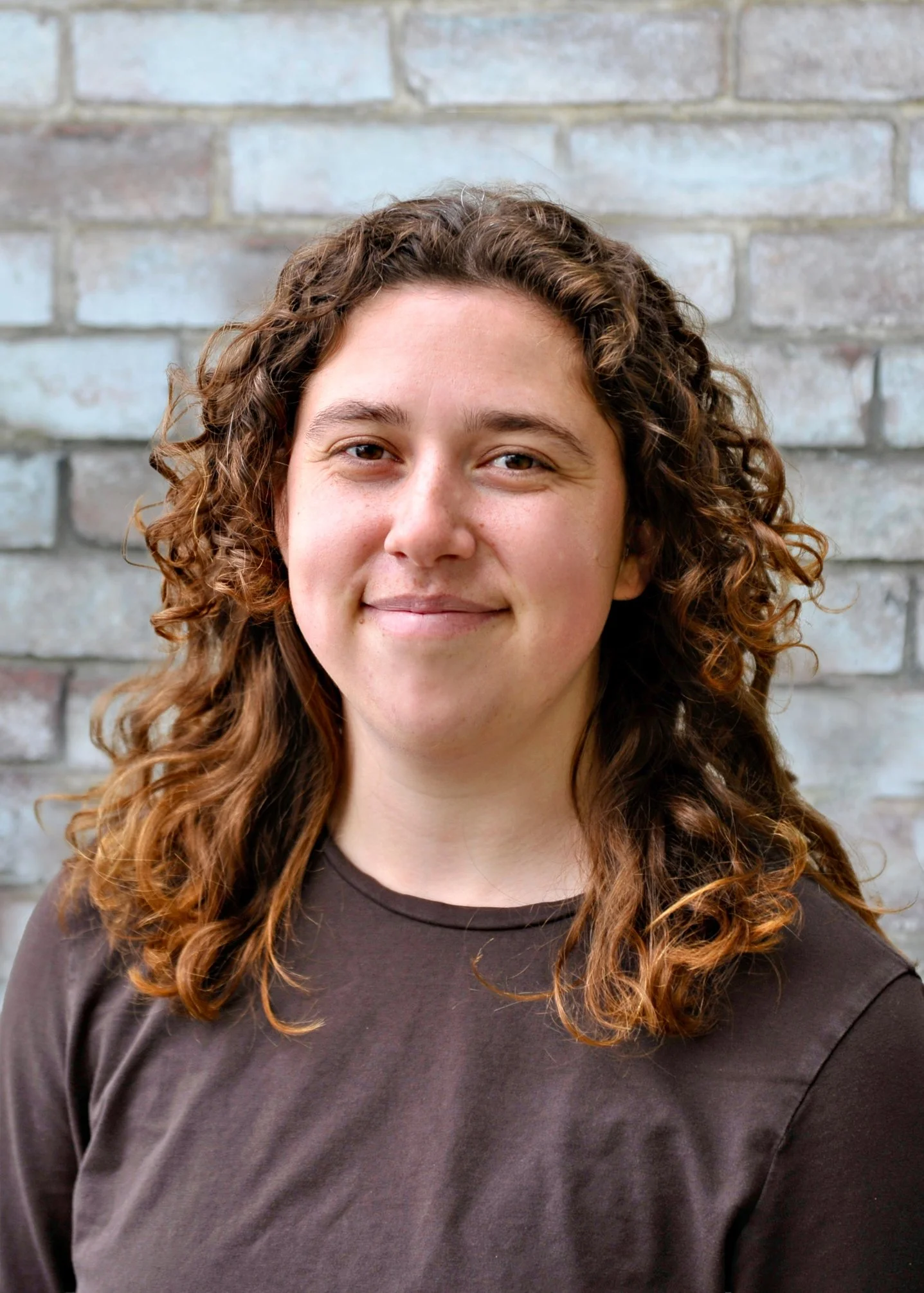 A young woman with curly brown hair, wearing a brown top, standing in front of a gray brick wall.