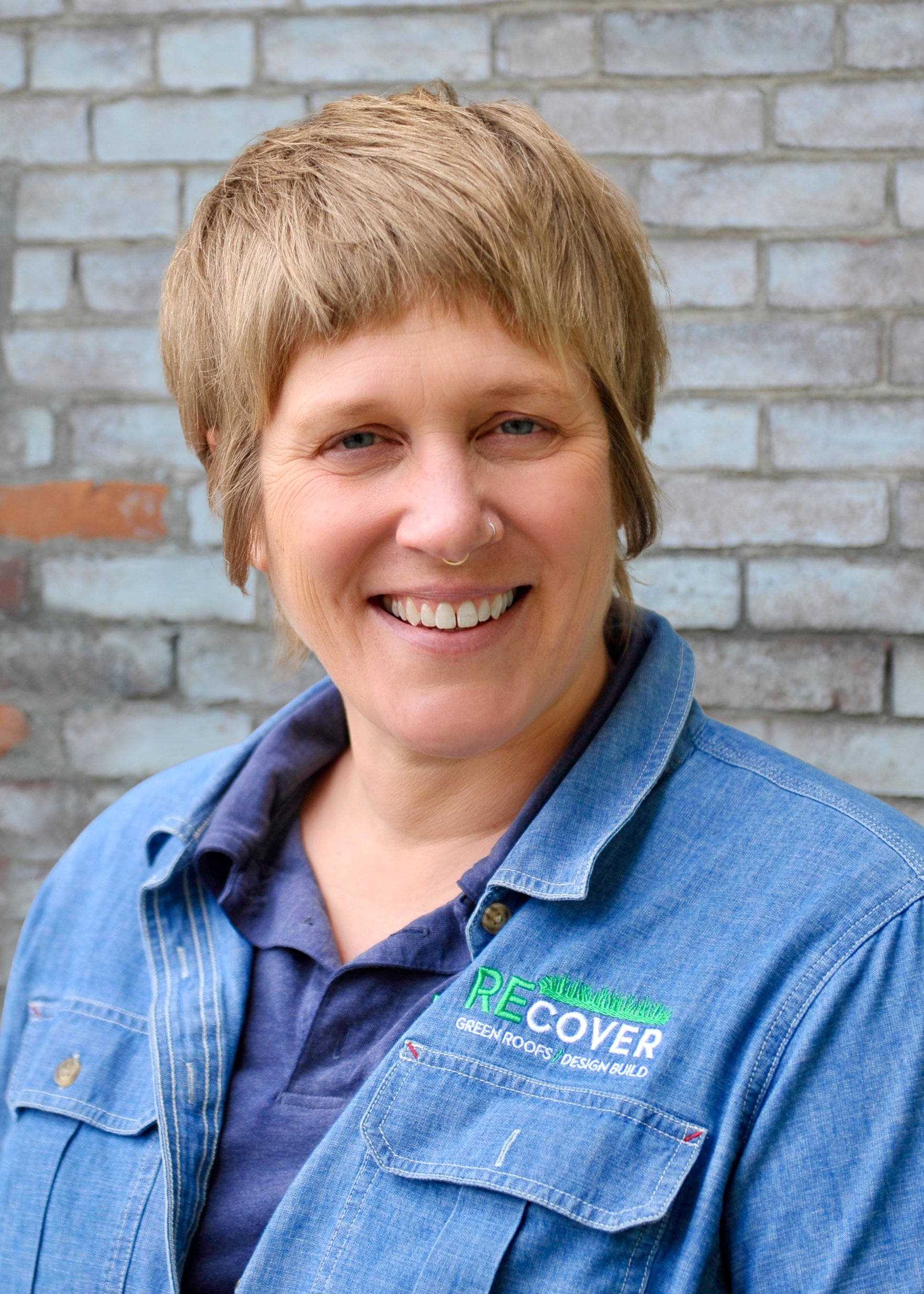 Close-up of a woman with short blonde hair, smiling, wearing a blue denim shirt with aGreener logo embroidered on it, standing in front of a brick wall.
