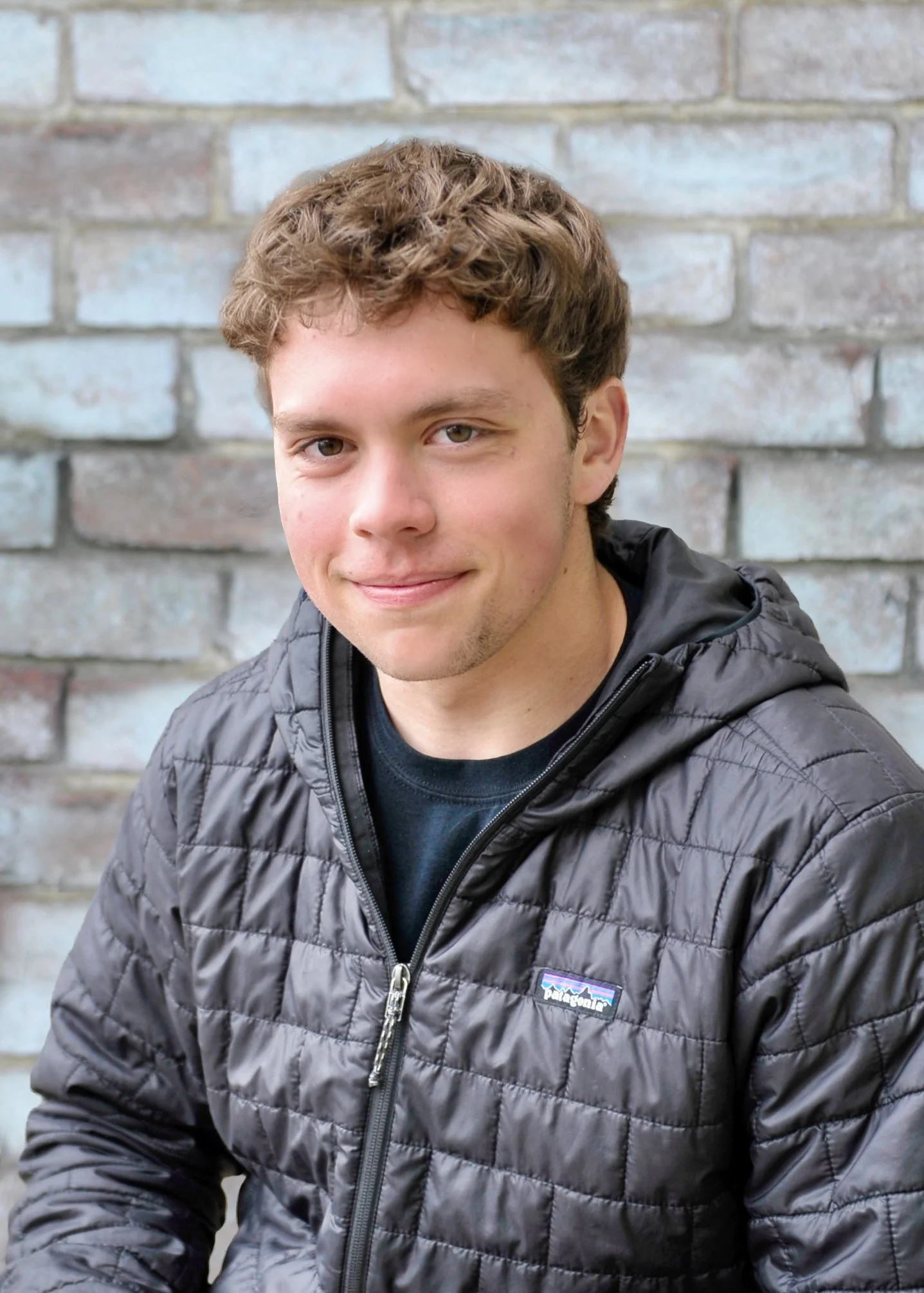 Close-up portrait of a young man with curly brown hair, fair skin, and a slight smile, wearing a black Patagonia quilted jacket, sitting against a gray brick wall background.