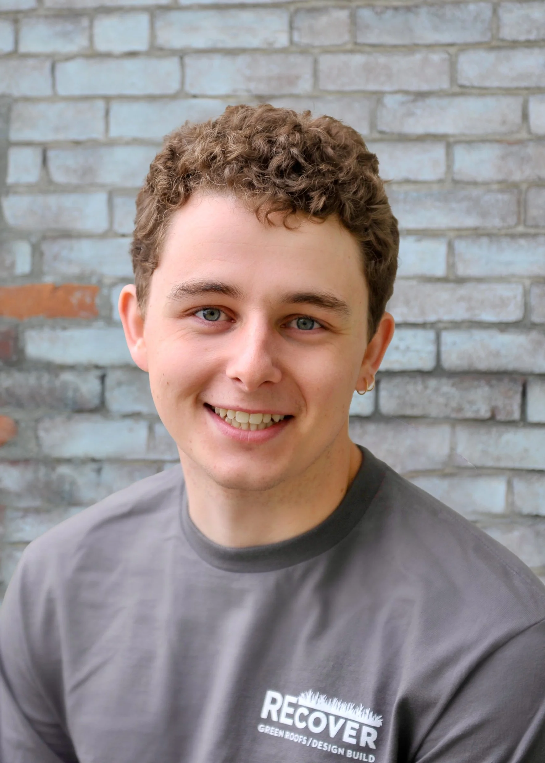 A young man with short, curly brown hair and light blue eyes smiling, wearing a gray T-shirt with a partial logo that reads 'RECOVER' over a background of a brick wall.