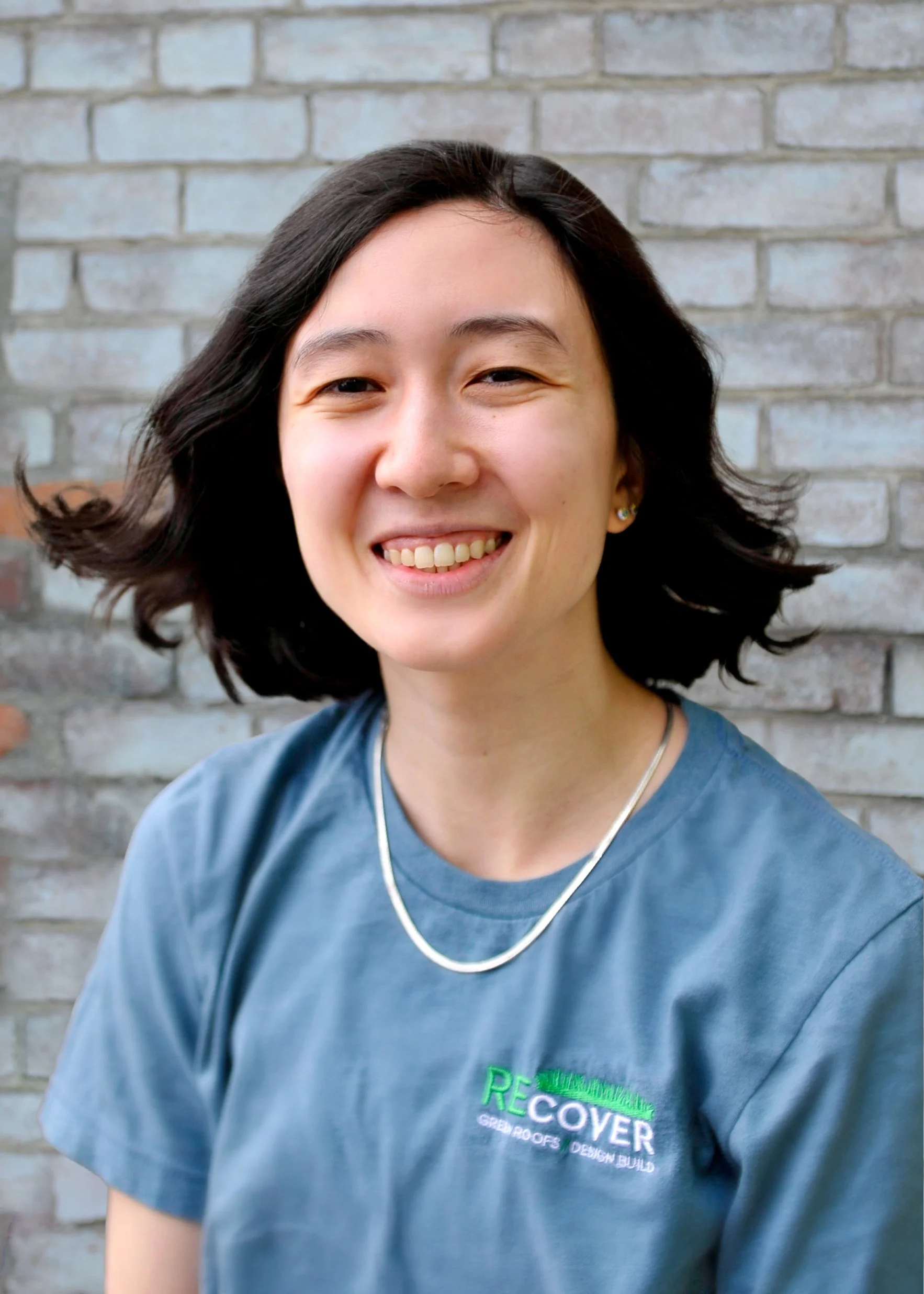 Close-up of a smiling woman with dark wavy hair and earrings, wearing a blue t-shirt with a logo, standing in front of a brick wall.