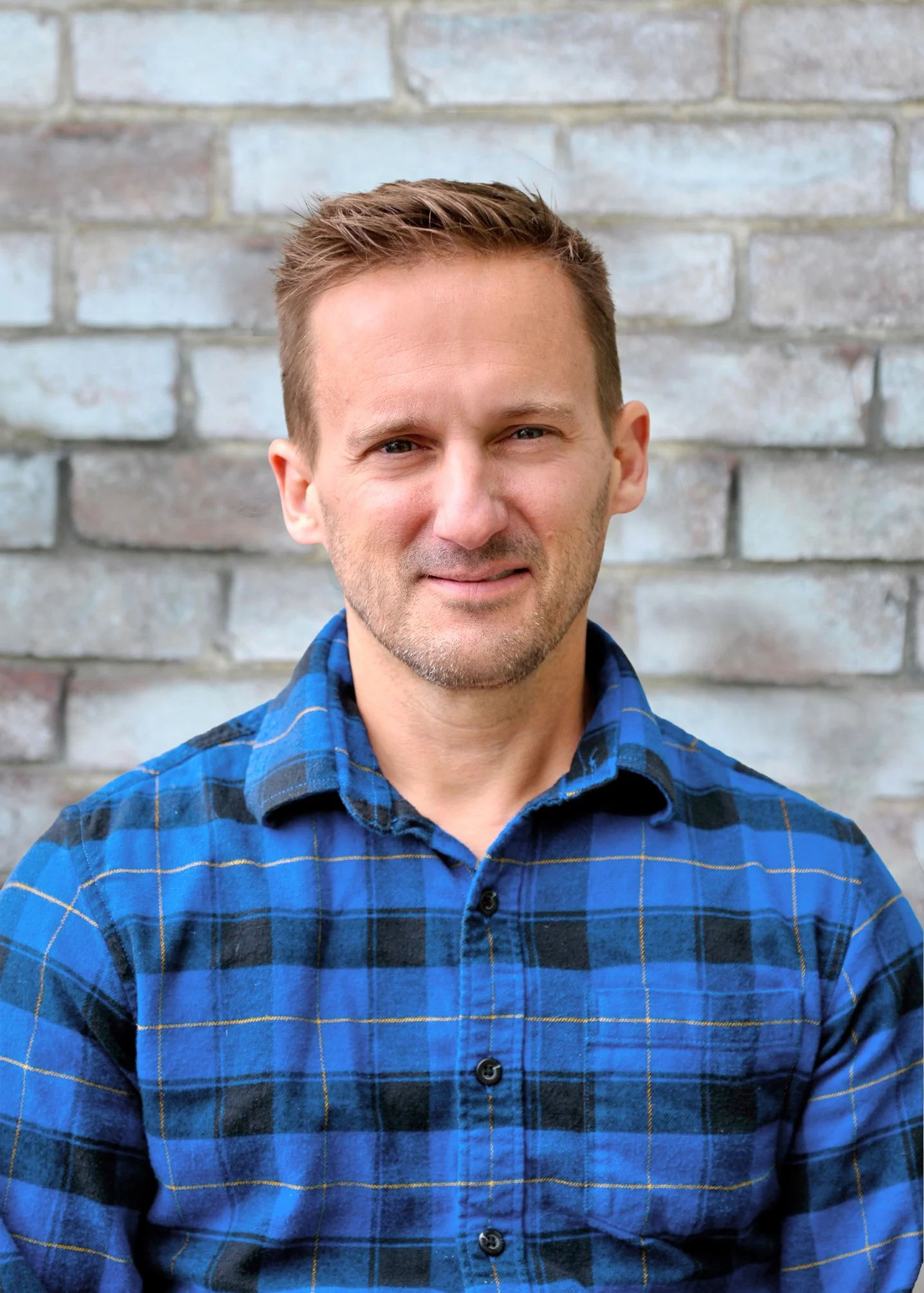 A man with short light brown hair and blue eyes smiling slightly, wearing a blue plaid shirt, standing in front of a brick wall.