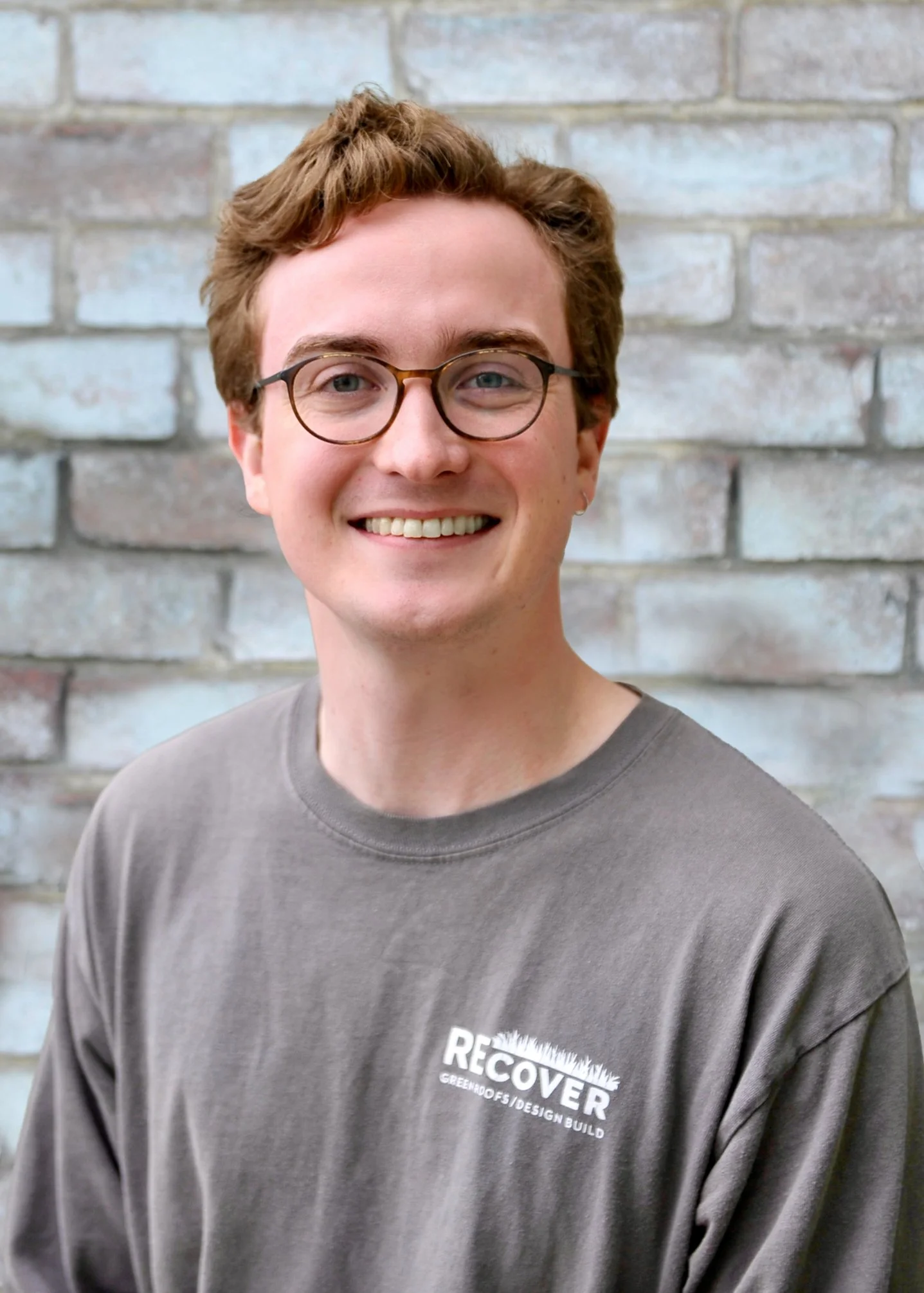 Young man with light brown hair, glasses, and a gray T-shirt smiling in front of a brick wall.
