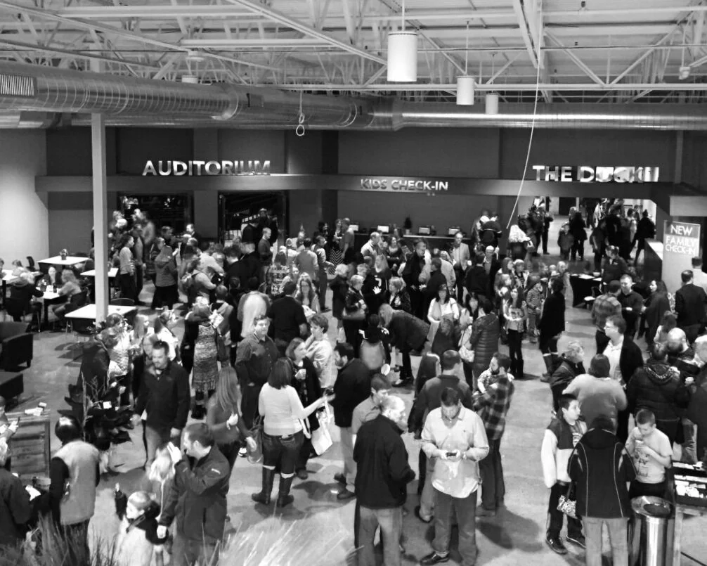 Black-and-white photo of community members socializing and gathering in a modern lobby hallway outside an auditorium.