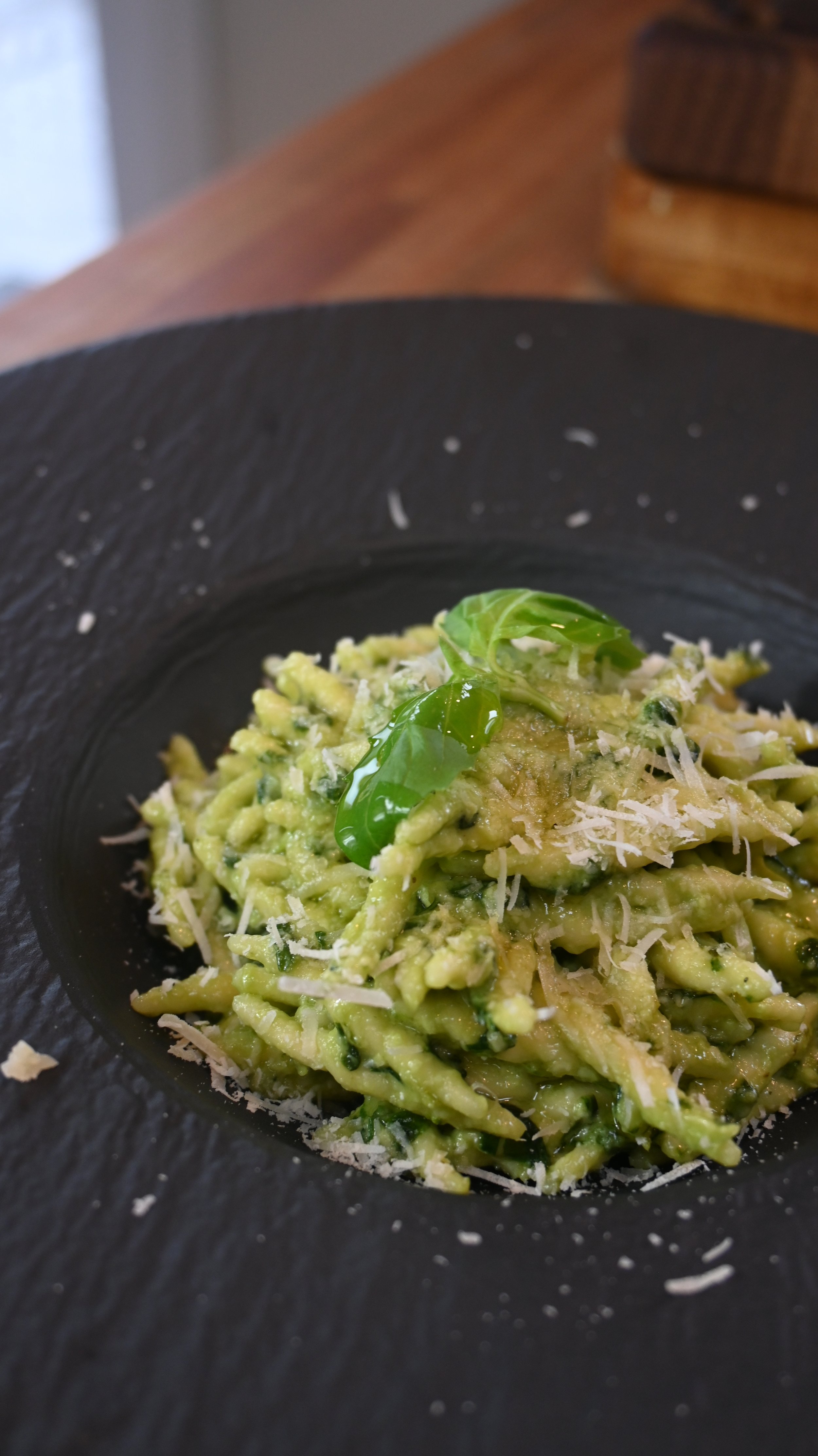 Close-up of a plate of green pesto pasta garnished with basil and grated cheese, served in a black bowl.