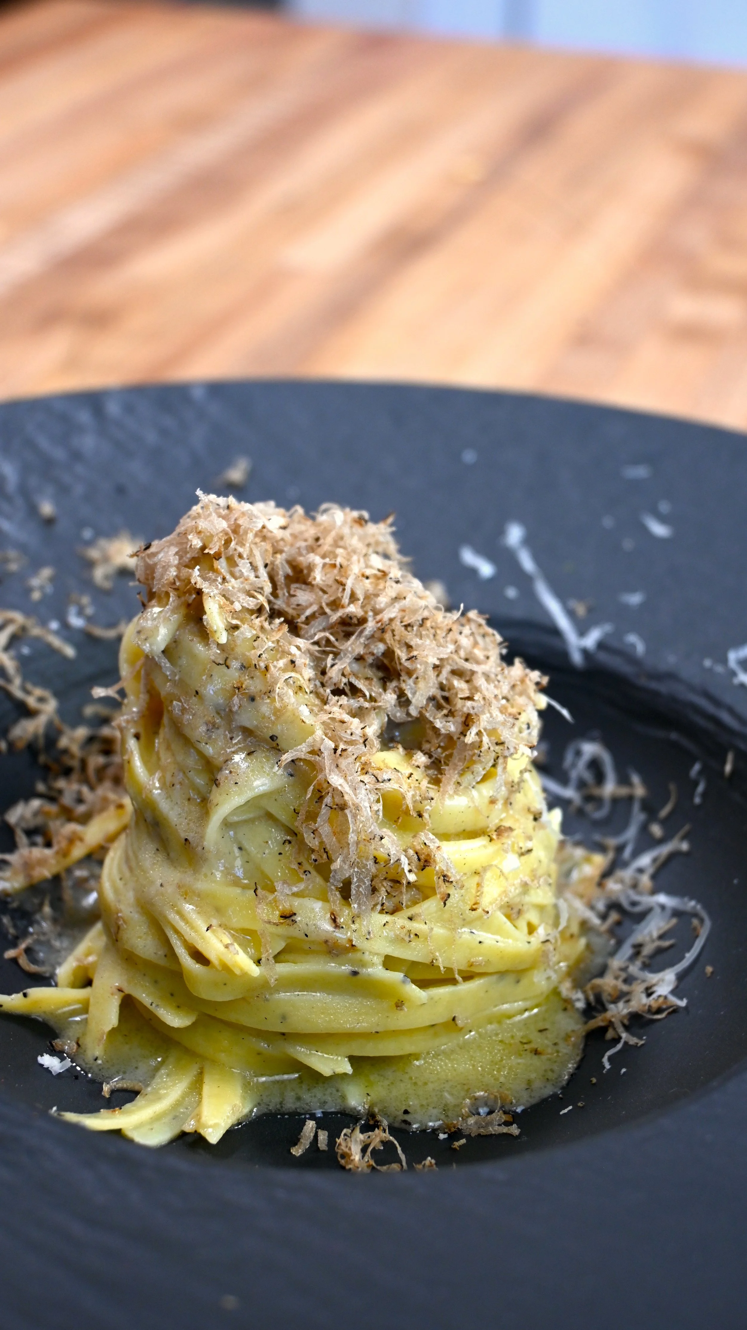 A plate of pasta topped with shredded cheese and bonito flakes on a black plate, with a wooden table in the background.