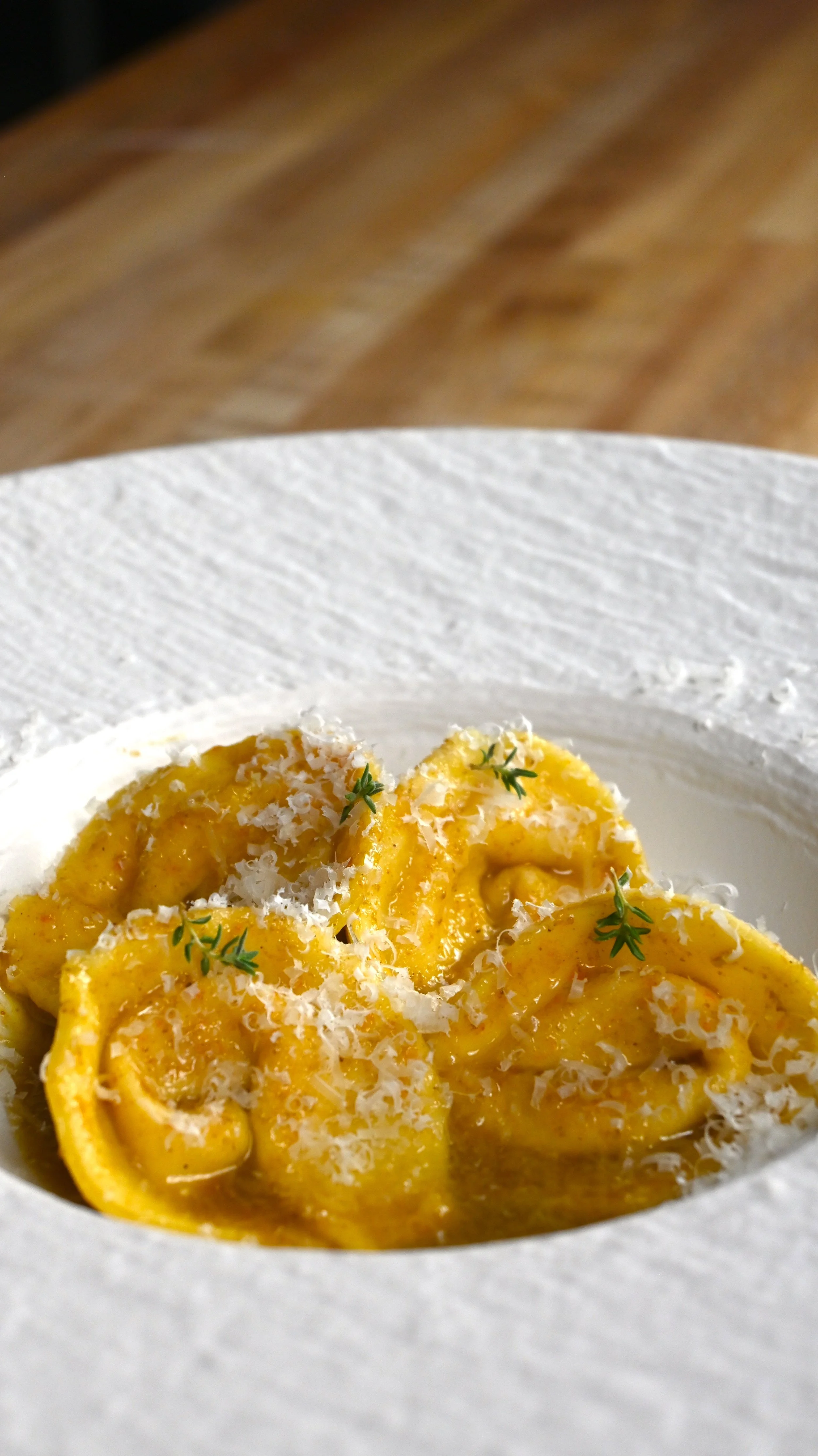 Close-up of a white bowl of ravioli pasta topped with grated cheese and garnished with small green herbs, set on a wooden surface.