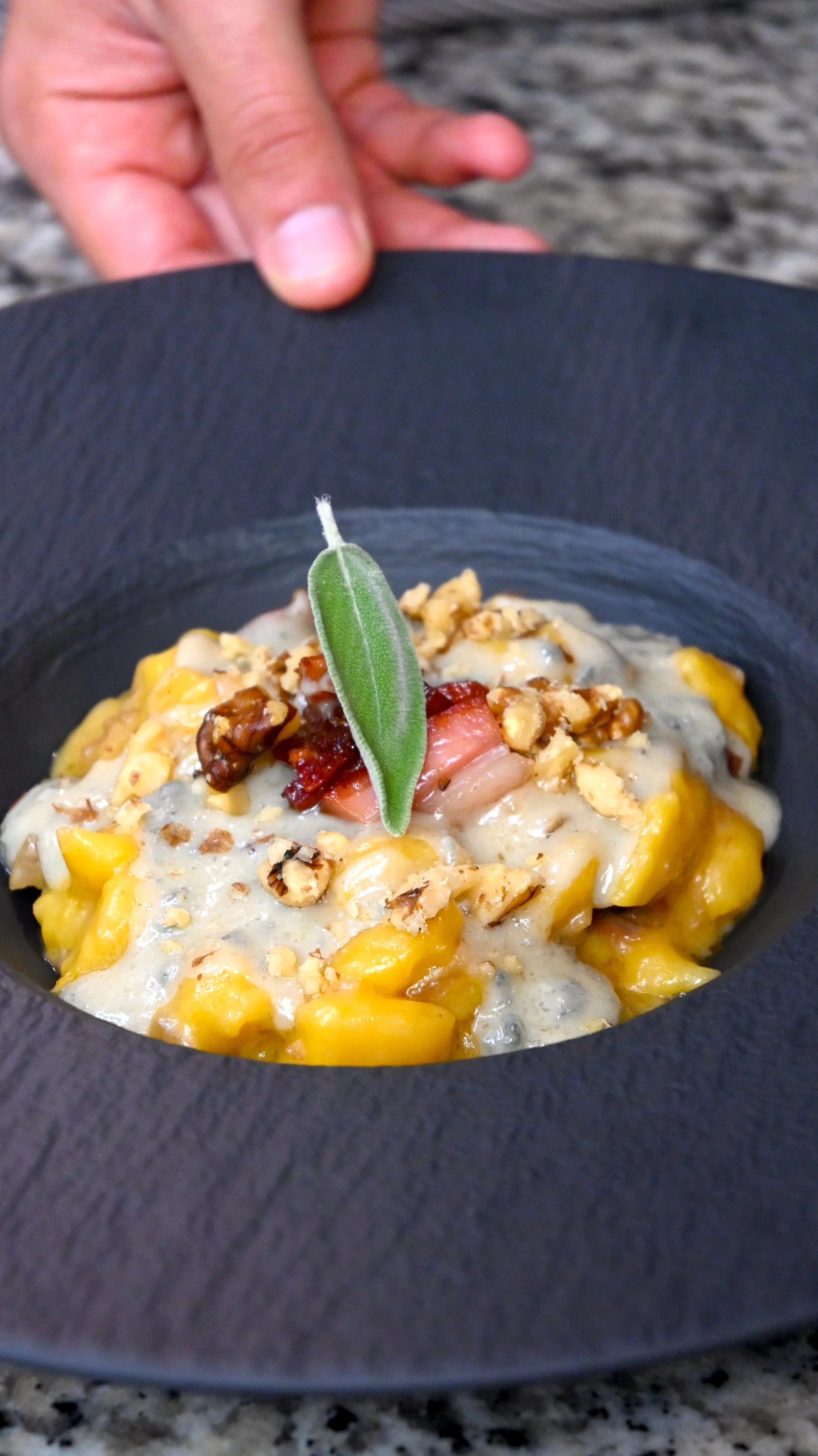 A bowl of yellow gnocchi topped with melted cheese, chopped nuts, bacon, and a sage leaf, held over a dark plate with a blurred background.