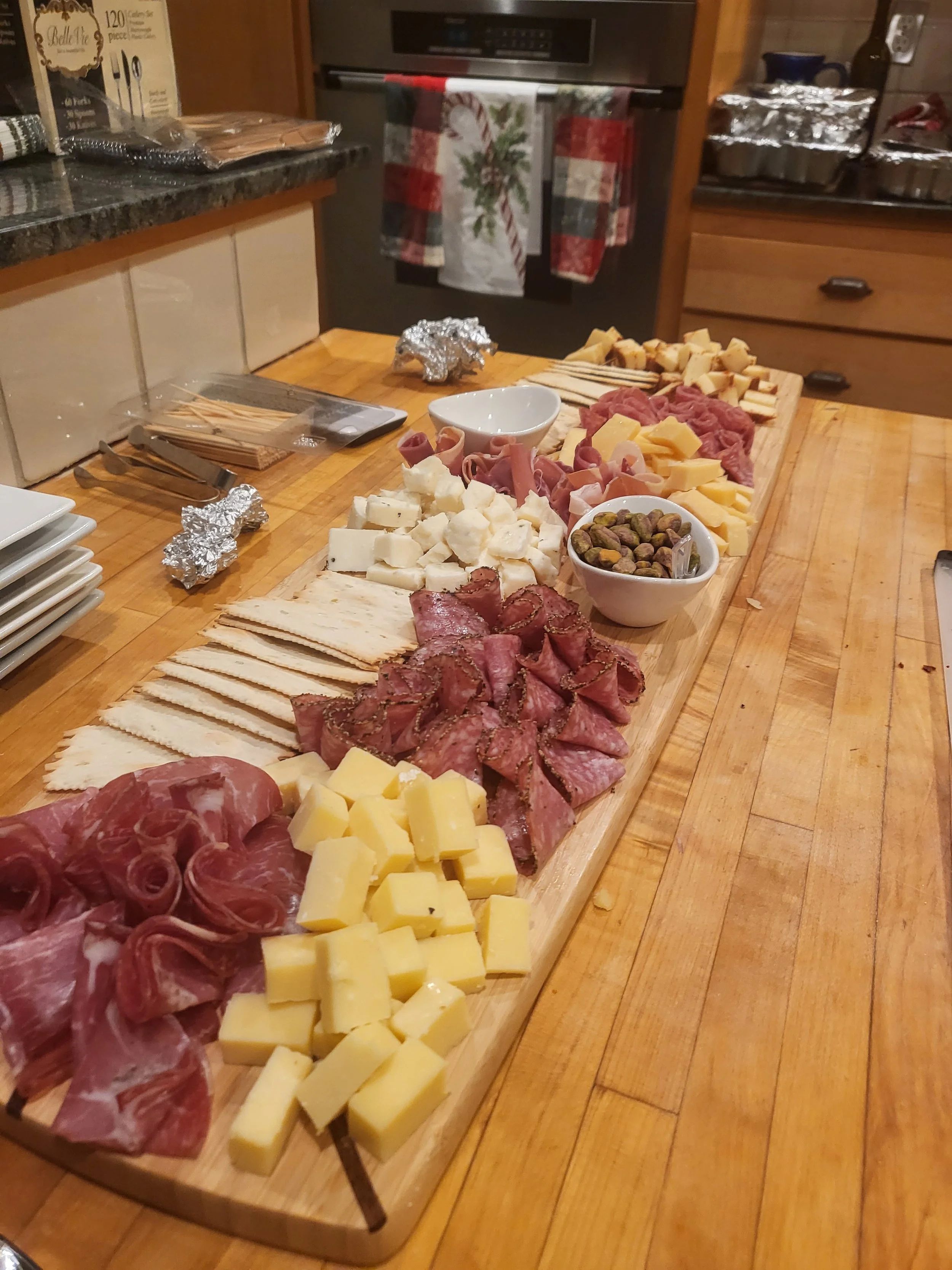 Charcuterie board with assorted cheeses, cured meats, crackers, and bowls of pistachios on a wooden table in a kitchen setting.