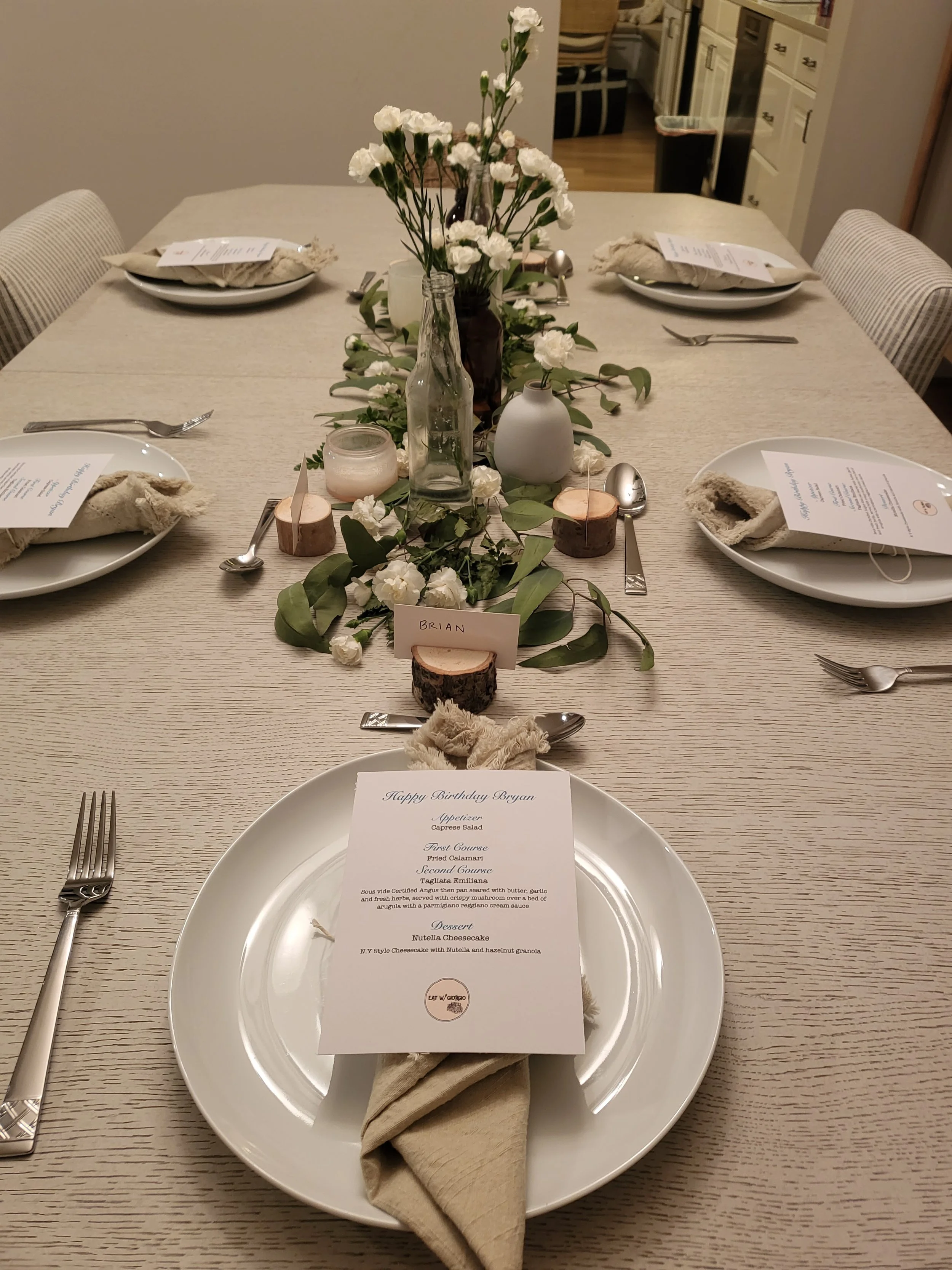 A dining table set for a birthday celebration with plates, cutlery, napkins, and a detailed menu card. The centerpiece features white flowers, greenery, and candle holders, with a name card reading 'Brian'.