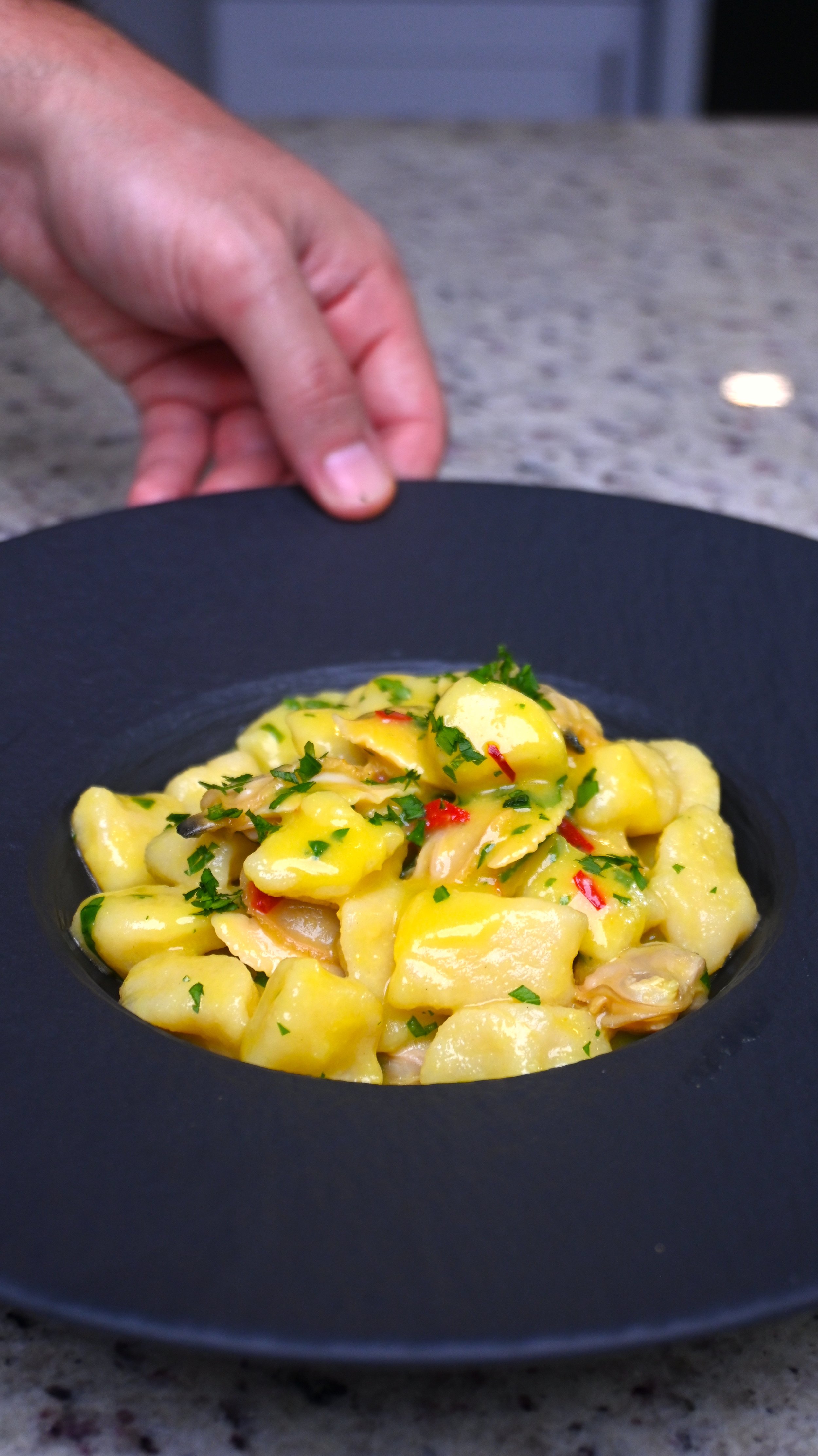 A black plate of yellow gnocchi with herbs and chopped red peppers on a granite countertop.