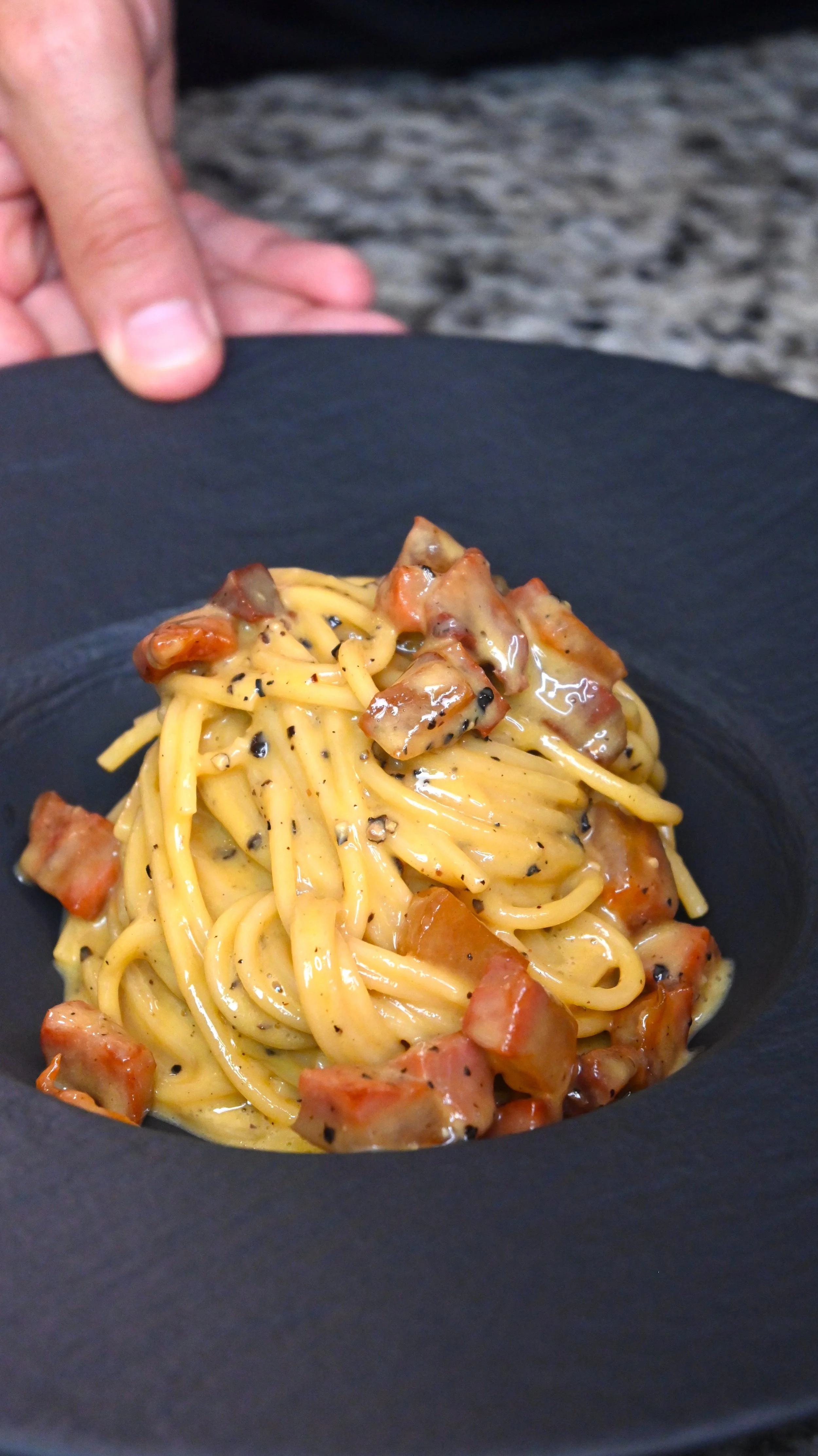 A plate of creamy spaghetti with bacon bits and black pepper on a black plate.