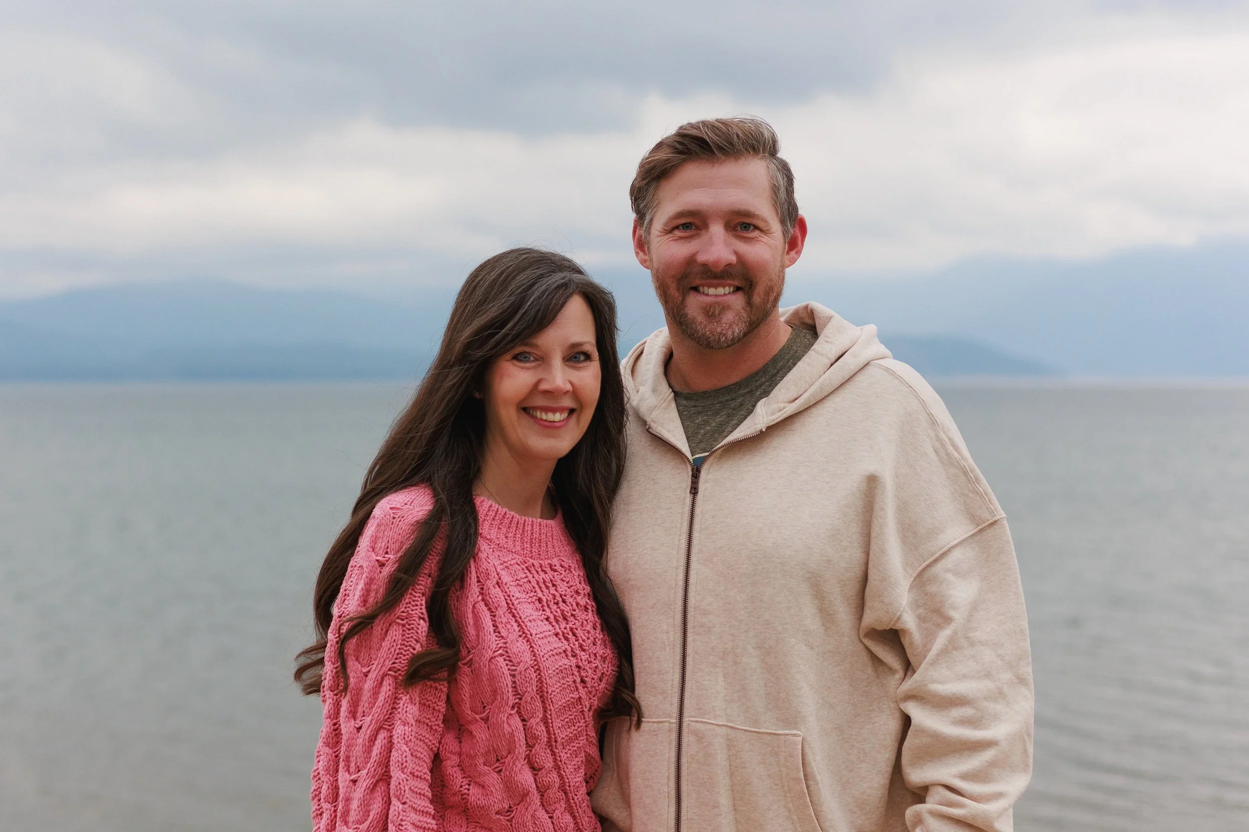 A smiling couple standing on a beach with water and cloudy sky in the background.