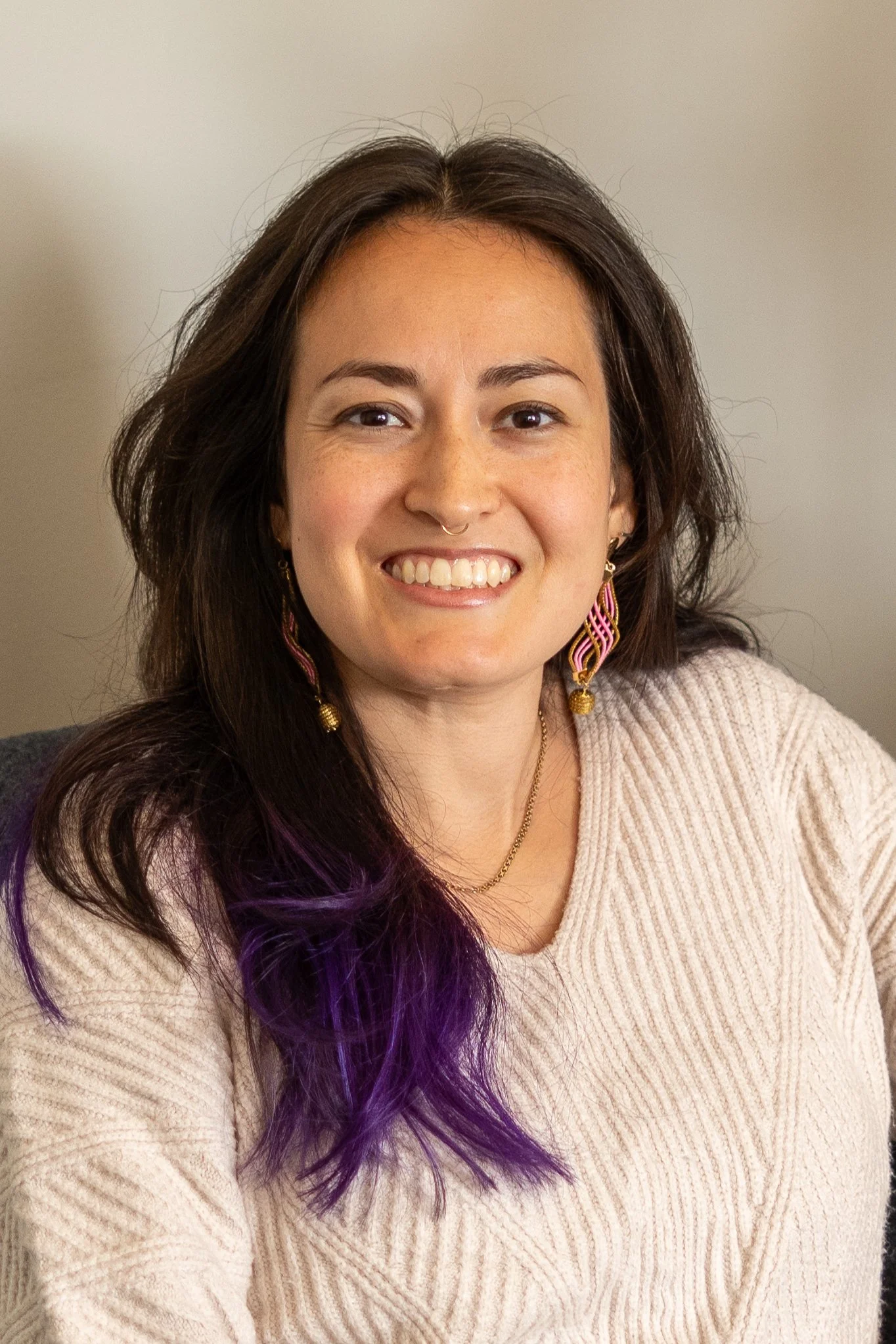 Portrait of a woman with long dark hair, some purple highlights, earrings, and a beige sweater, smiling at the camera.